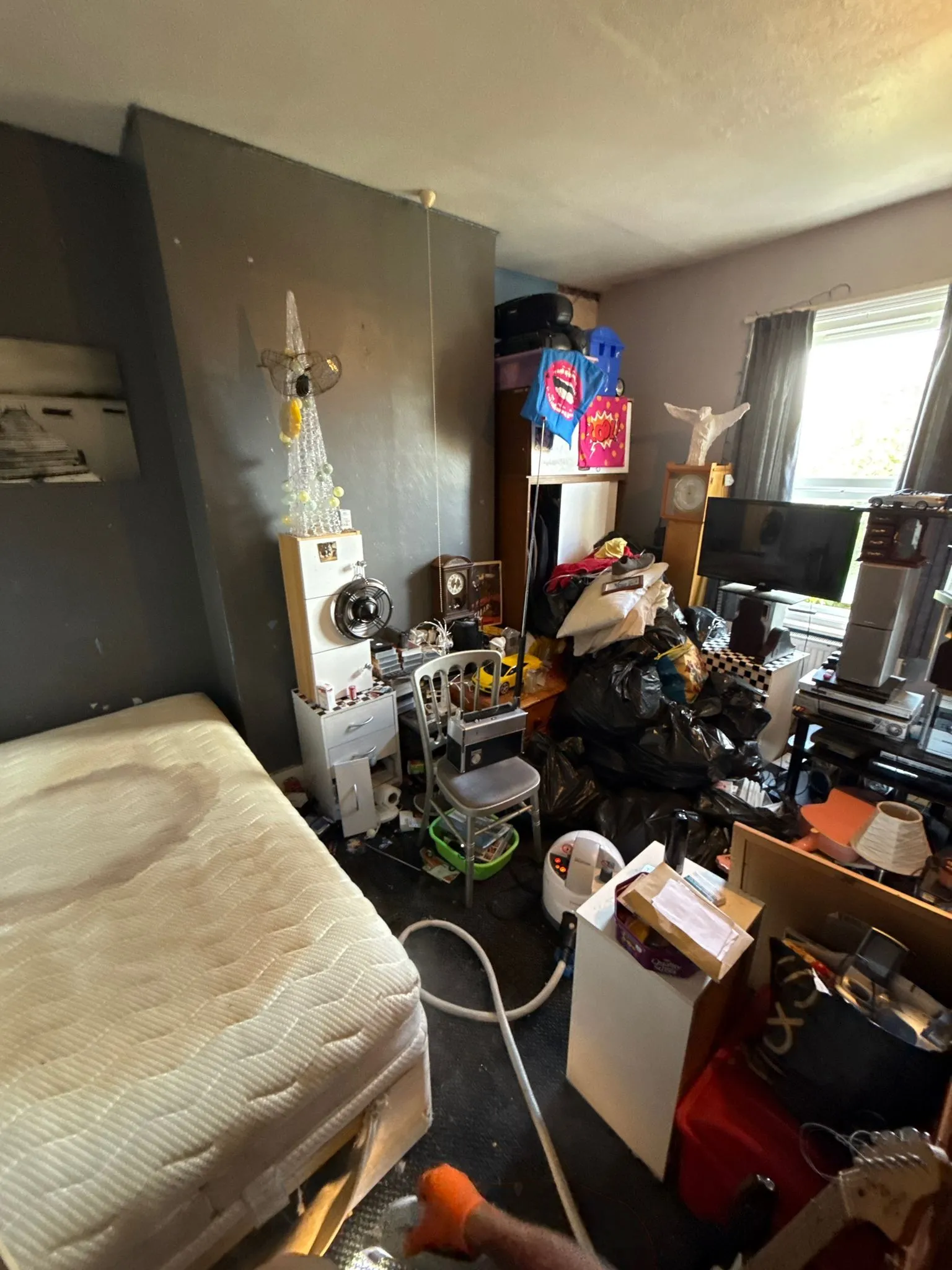 A highly cluttered and messy room with a bed, black bin bags, and general disarray, suggesting a need for comprehensive pest control services.
