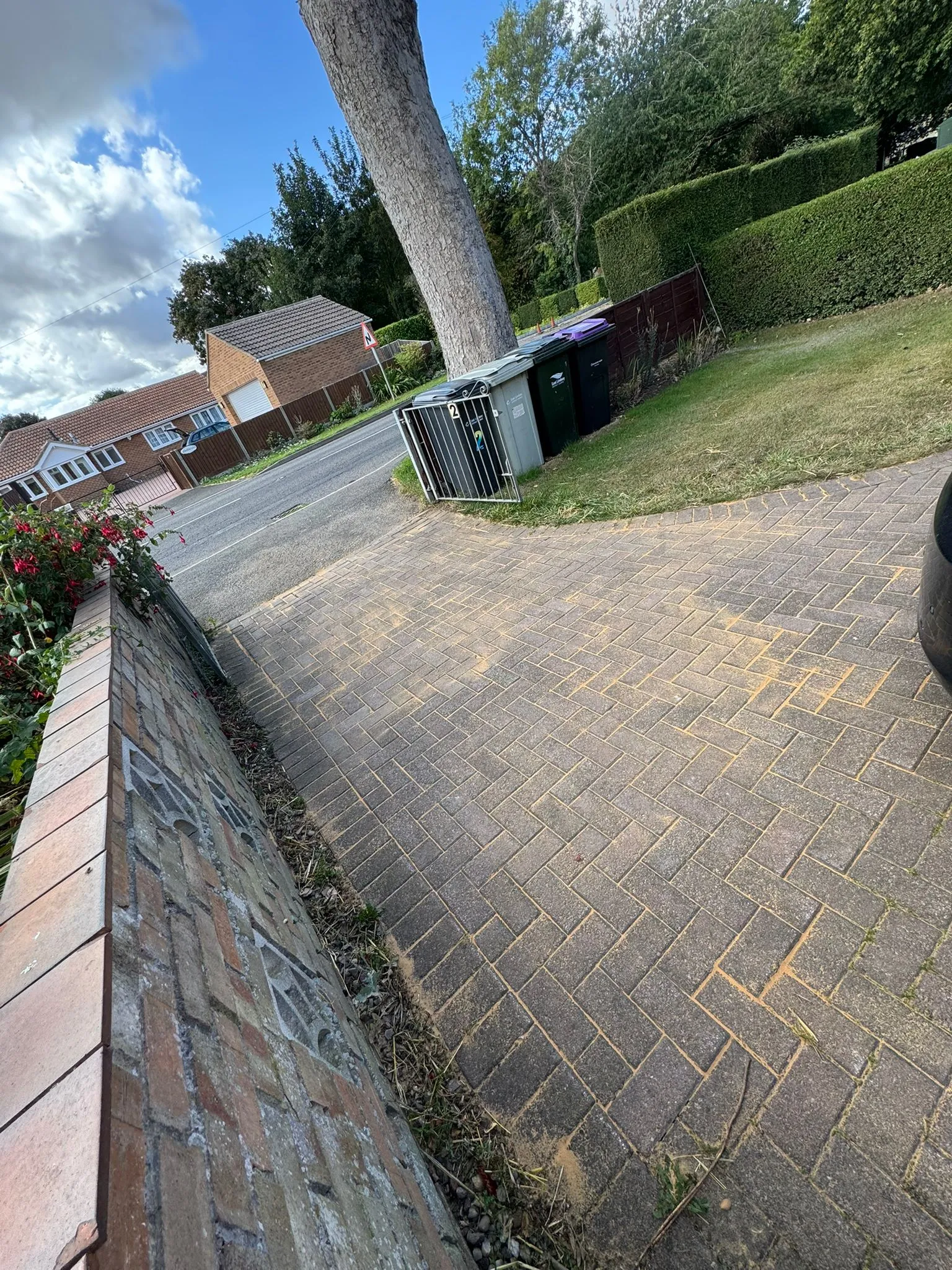 A block paved driveway with sand-filled joints, indicating recent installation or maintenance, adjacent to a brick wall and flowering plants.