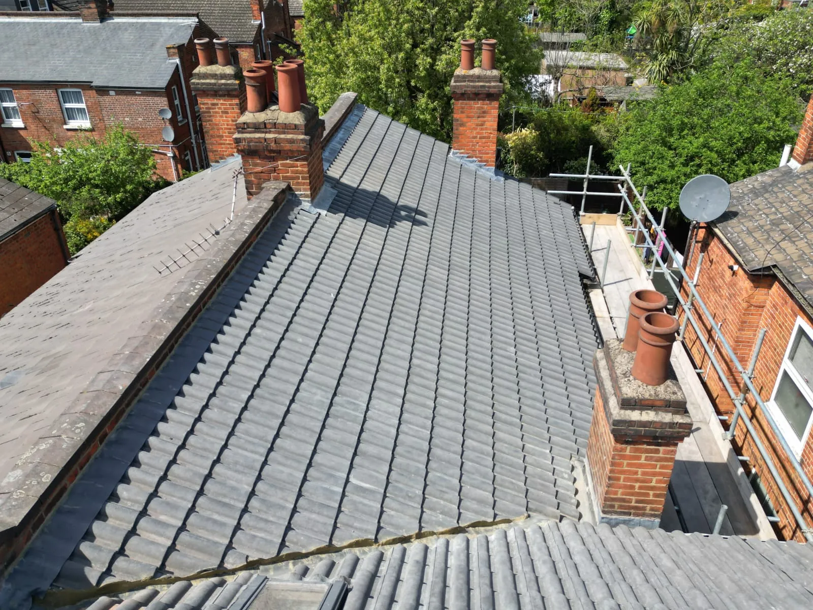 A close-up view of a residential roof featuring newly installed grey tiles next to older roofing materials, with prominent brick chimneys and scaffolding, indicating a re-roofing or new roof installation project.