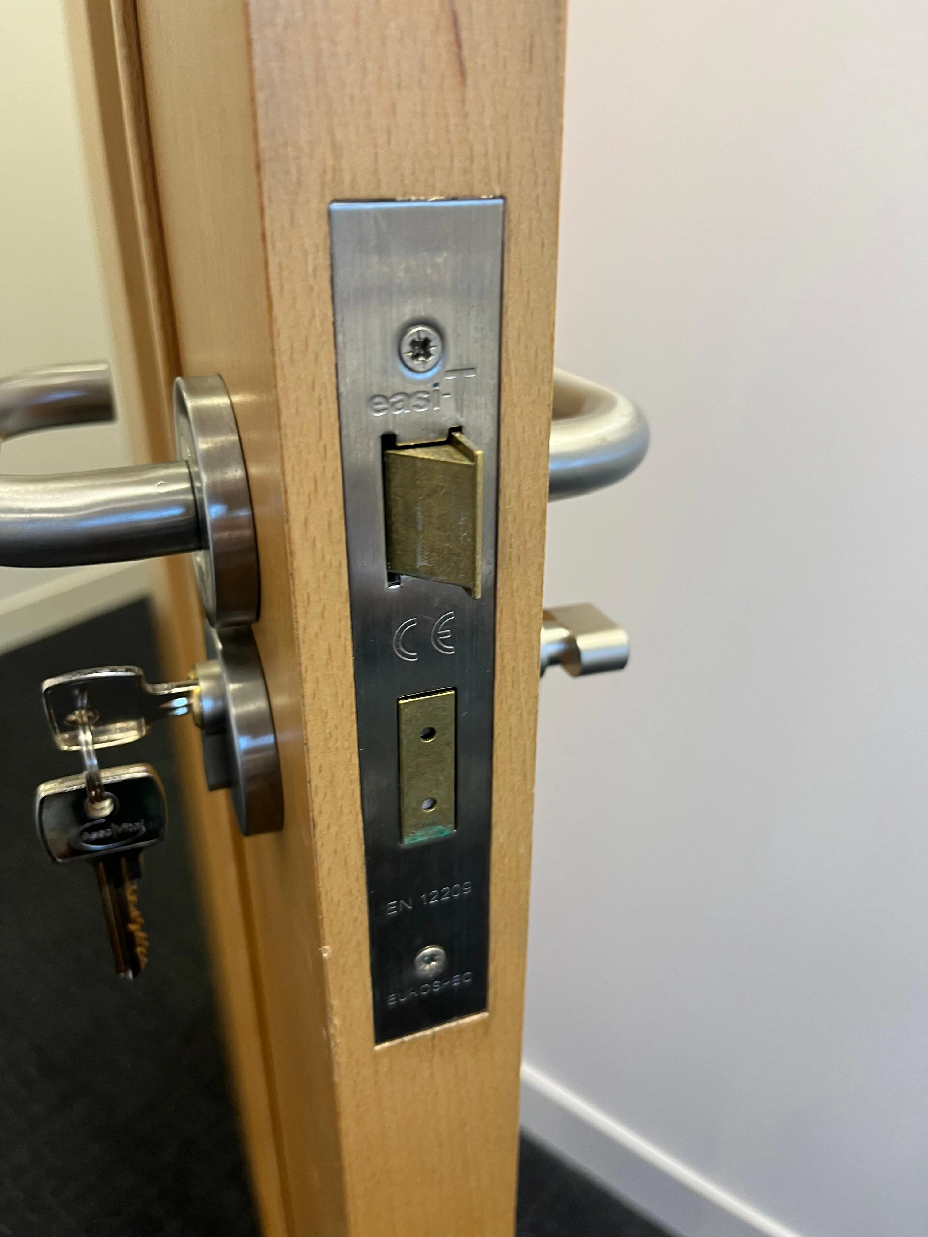 Keys inserted into a chrome mortice lock on a light wooden door with a matching handle, viewed from the side.