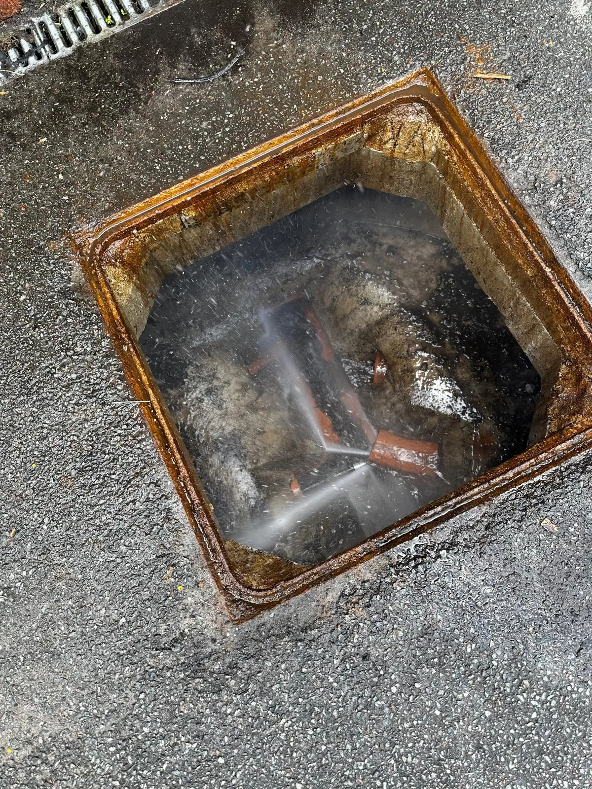 A high-pressure water jet actively cleaning a drain inside an open manhole, with water visibly spraying and pipes at the bottom.