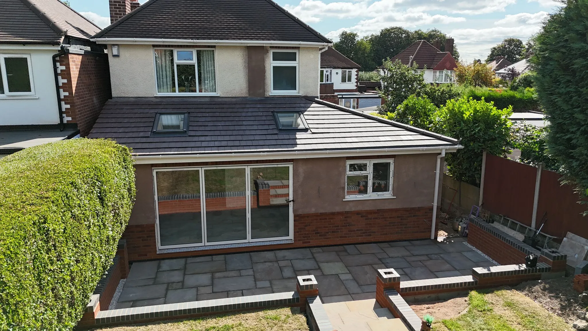 Exterior view of a newly built single-storey home extension featuring dark tiled roof, skylights, light brown render, bi-fold doors, and a grey paved patio.