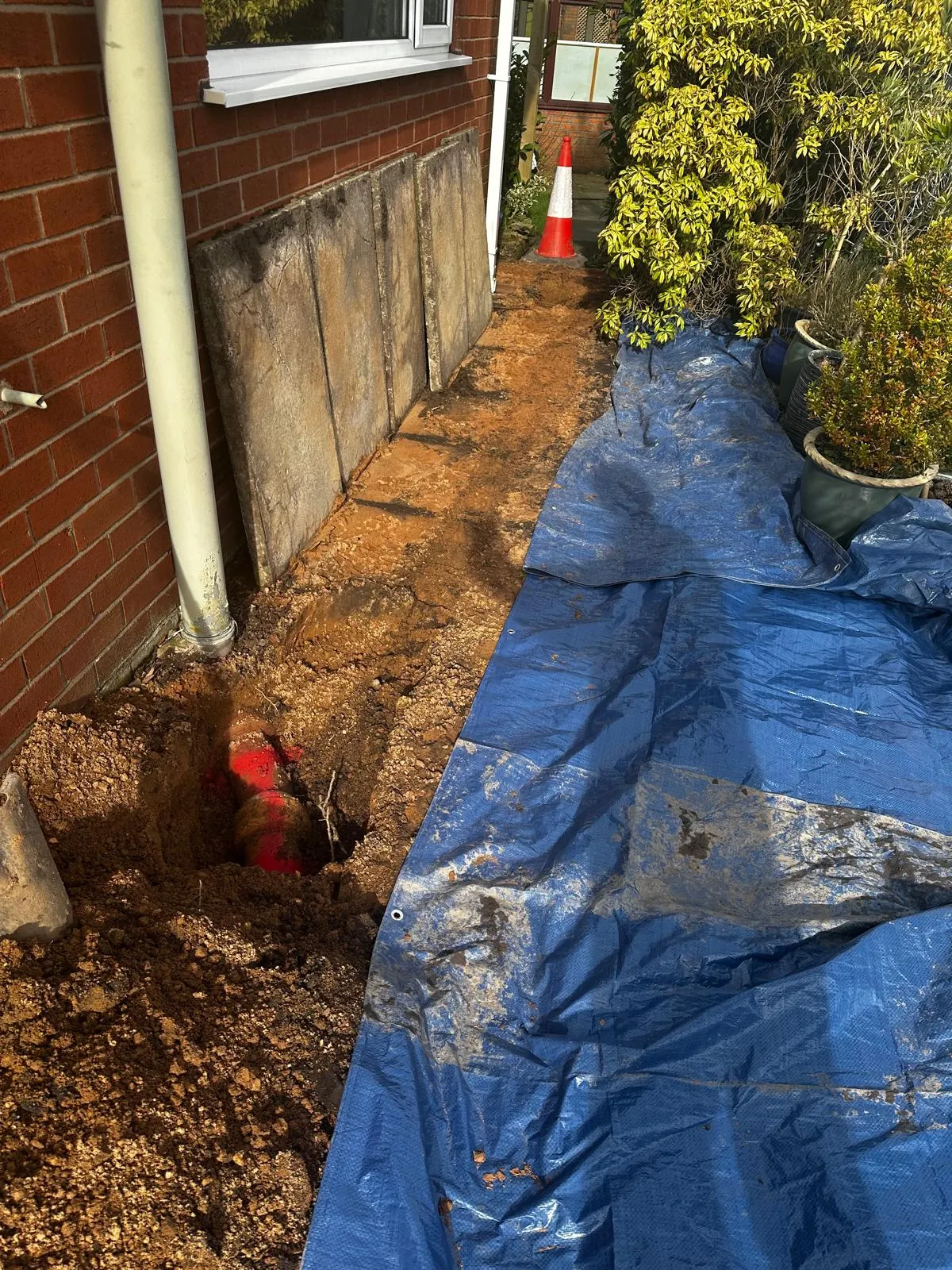 Excavation work showing exposed red drainage pipes next to a house, with a blue tarpaulin in the foreground, indicating a drainage repair or installation.