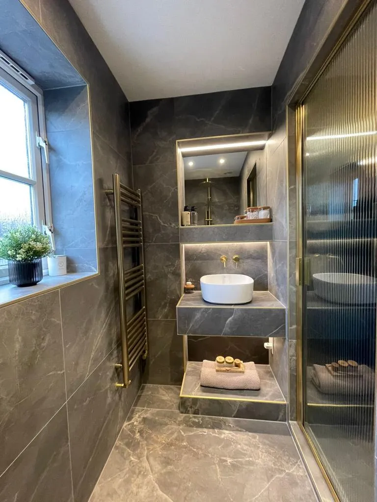 High-end luxury bathroom renovation in Plymouth by Sands & Vossen featuring dark marble tiling, gold brassware, and a fluted glass walk-in shower.