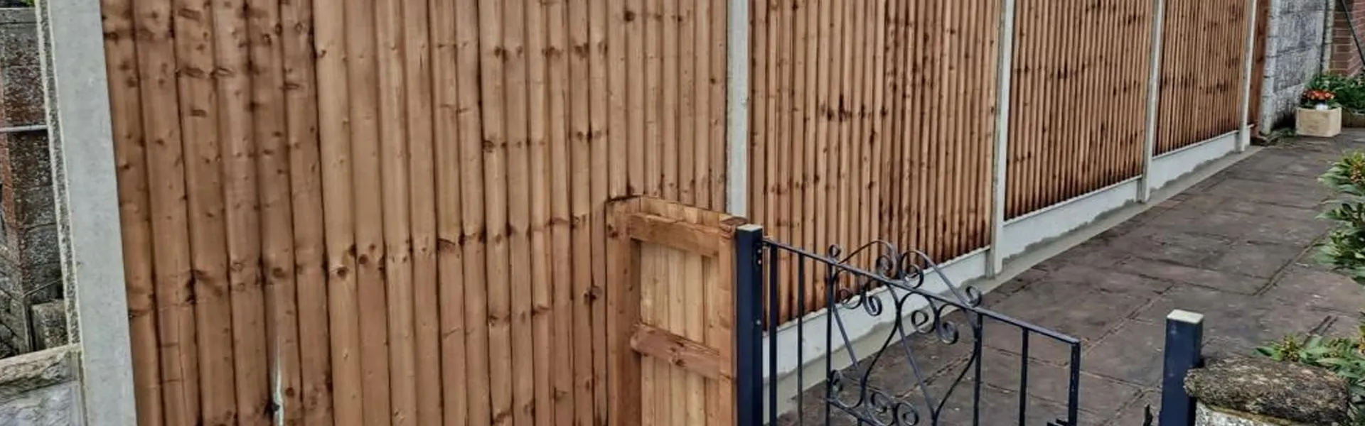 Newly installed wooden garden fence with concrete posts, an integrated wooden gate, and a black metal decorative gate on a paved path.