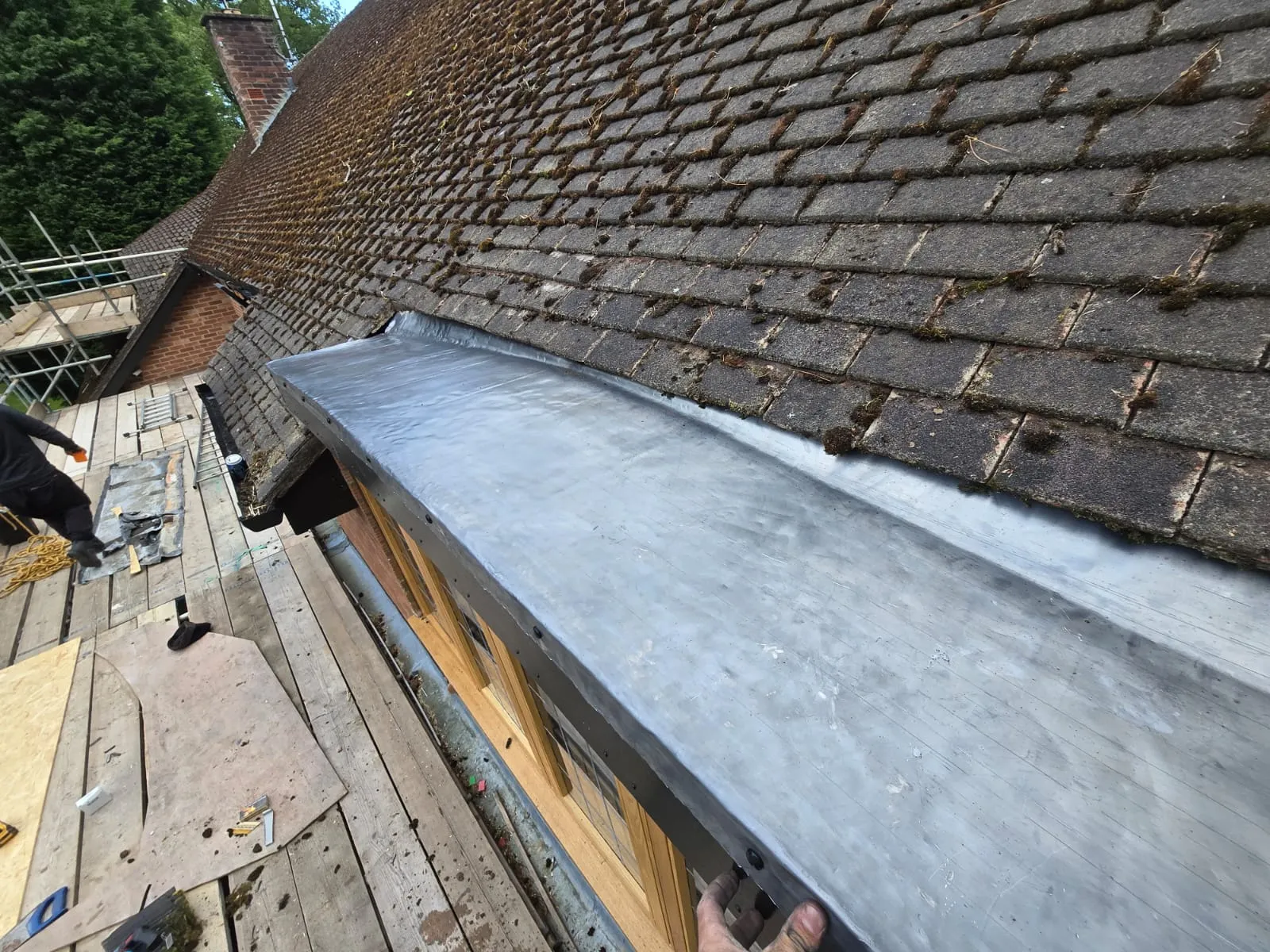 Newly installed traditional lead work on a roof, next to old, mossy roof tiles, with scaffolding and a roofer in the background.