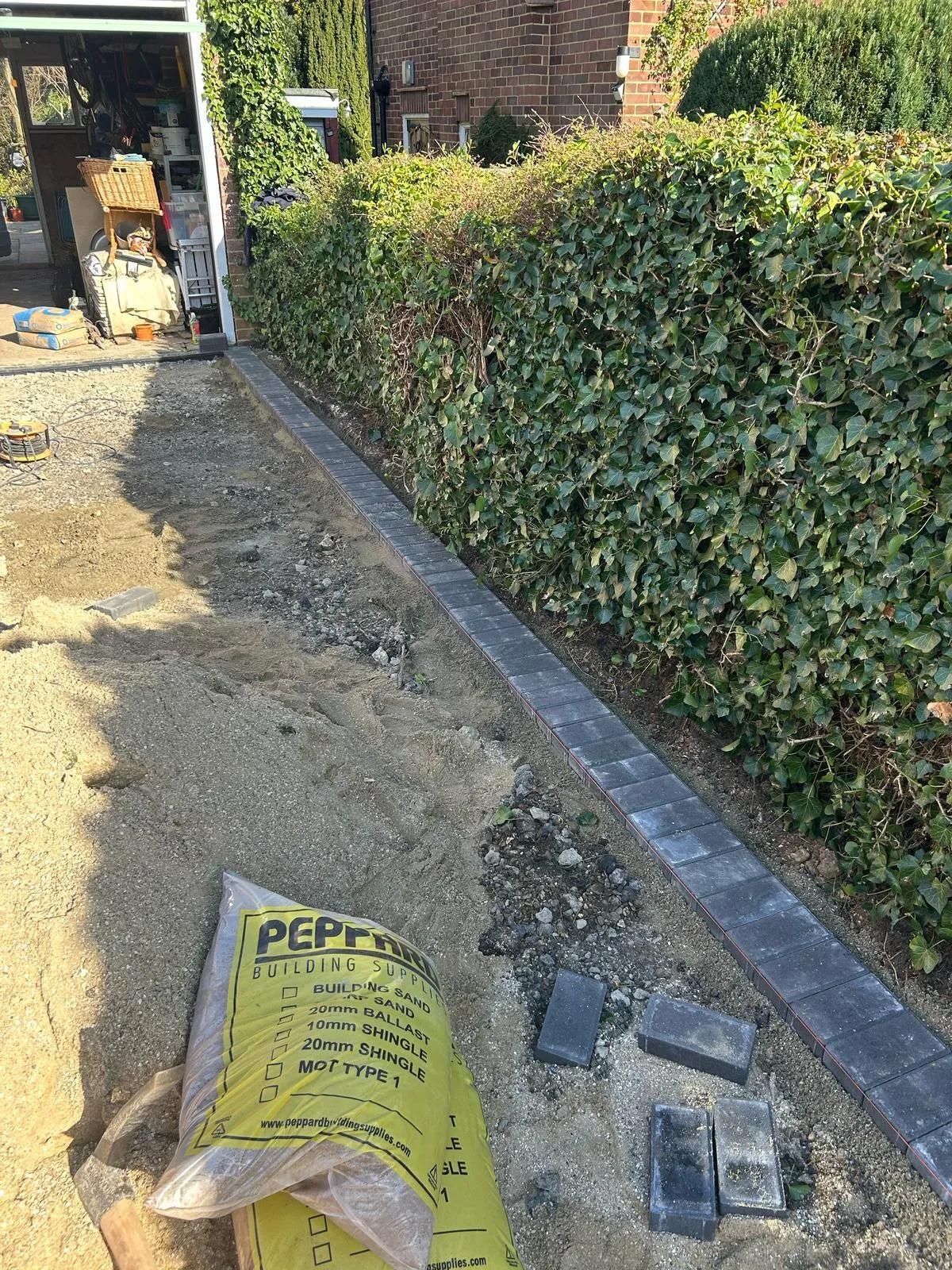 Work in progress on a new block paving driveway, showing grey paver edging installed next to a green hedge with sand and building supplies.