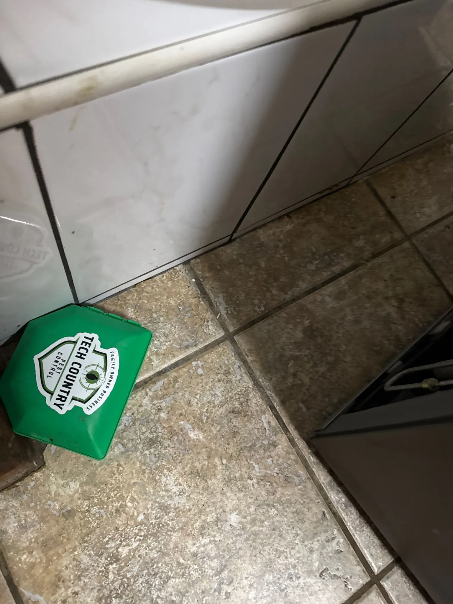 A green pest control bait station with the Tech Country Pest Control logo on a tiled floor.
