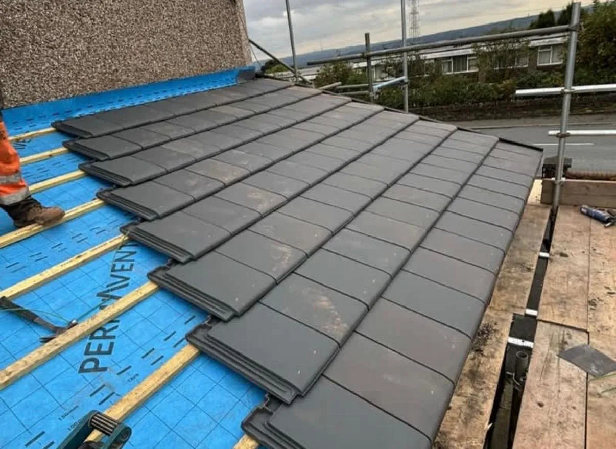 Roofer installing new grey tiles on a pitched roof with blue underlay and wooden battens in West Yorkshire.