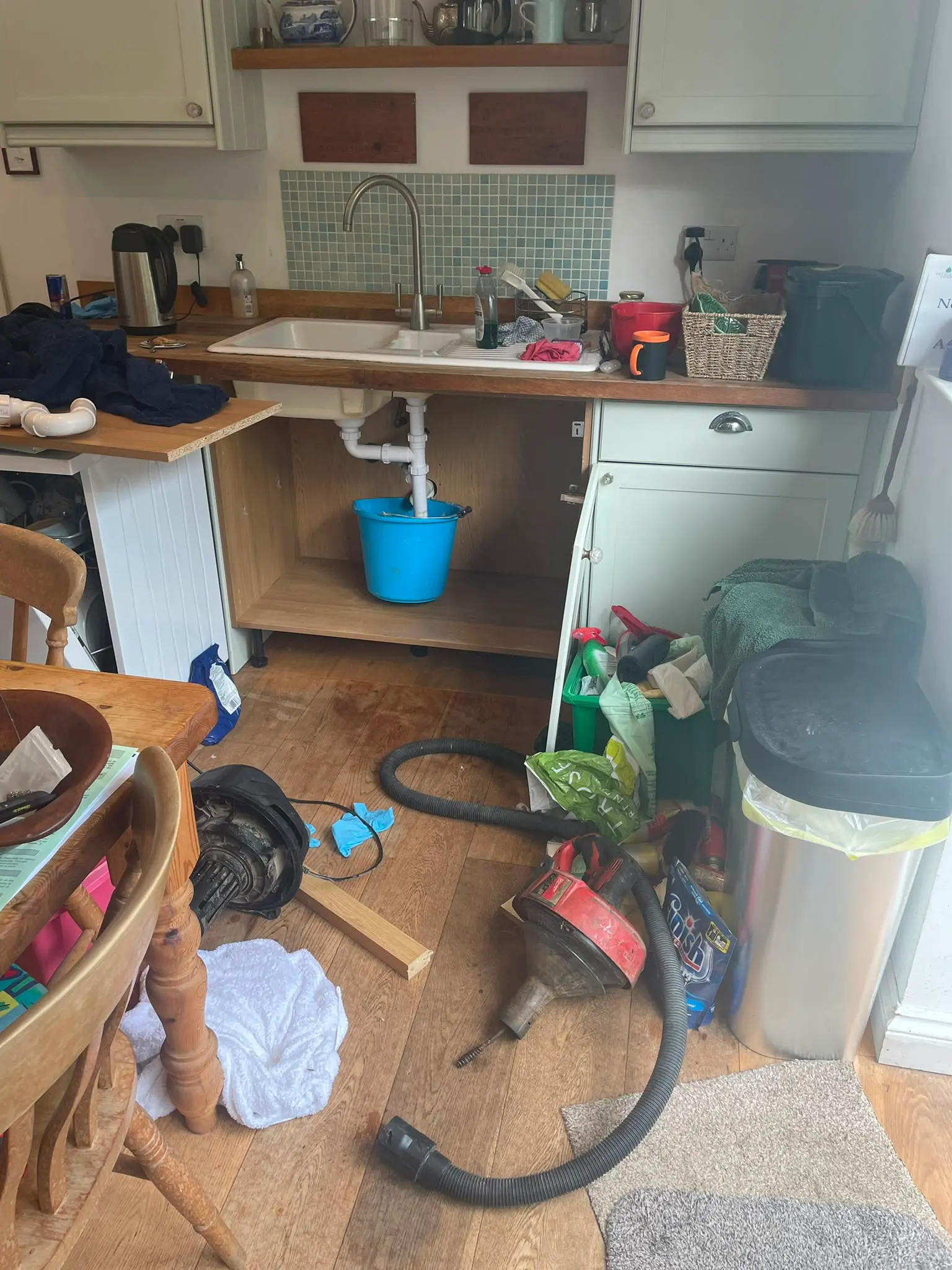 Plumbing tools including a drain auger and wet vacuum on a kitchen floor during a blocked sink repair