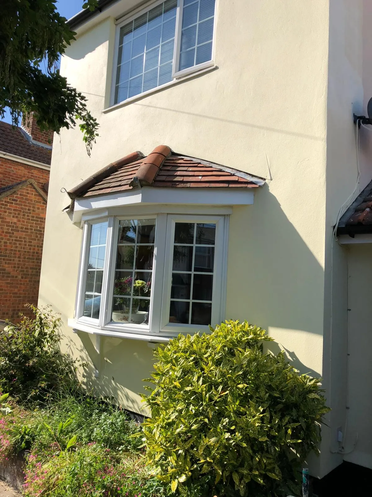 Exterior of a residential property with new cream render, white uPVC bay window, and upper casement window, bathed in sunlight.