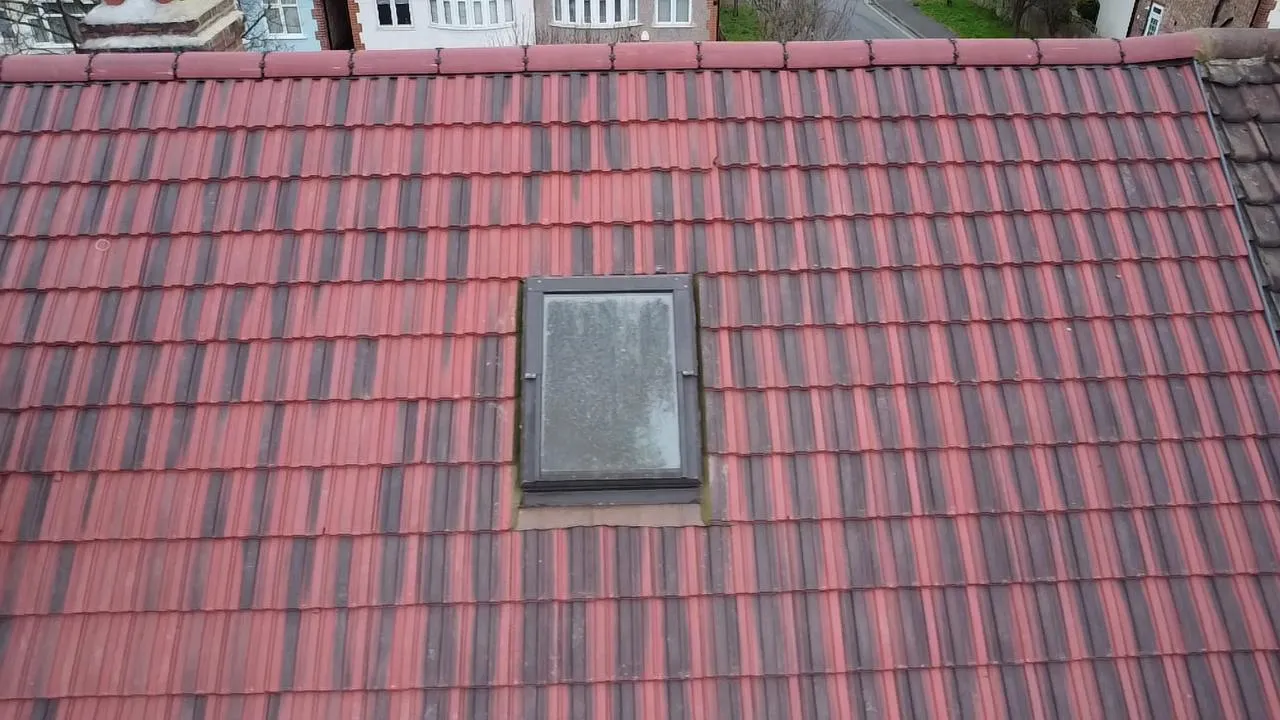 A close-up view of a tiled roof in shades of red and dark grey, with a rectangular Velux roof window installed in the centre.