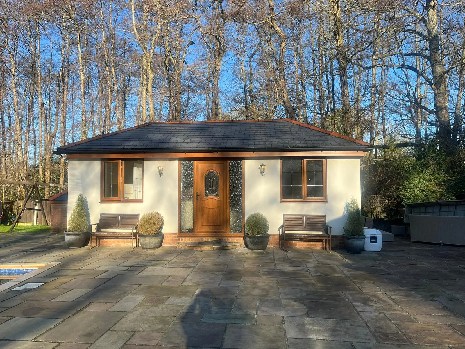 A close-up view of a recently installed dark grey tiled roof on a detached outbuilding with light render walls, brown windows, and a wooden door, showing the roofline and gutters.