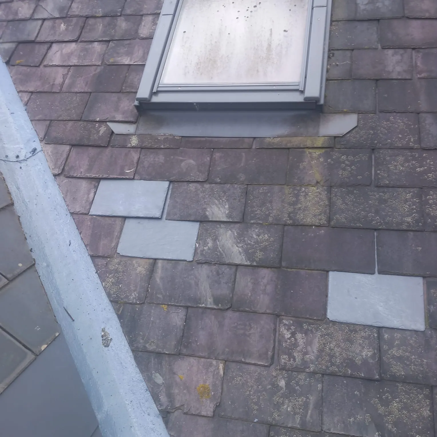 A pitched roof with a skylight, lead valley, and a mix of old, mossy slate tiles and new, lighter grey replacement tiles, indicating recent repairs.