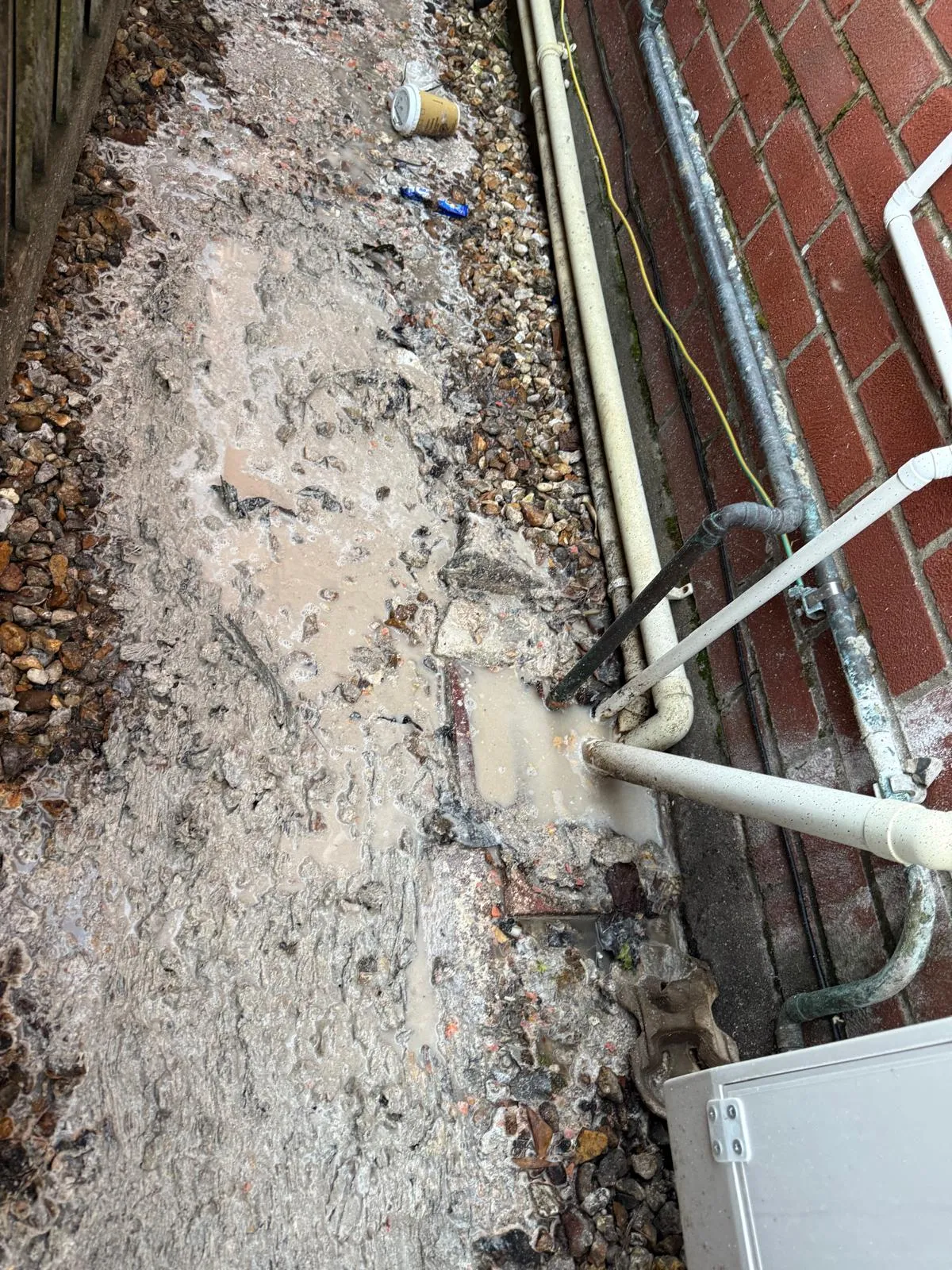 An image showing dirty, muddy water overflowing from a blocked external drain onto a gravel path, with pipes visible against a brick wall.