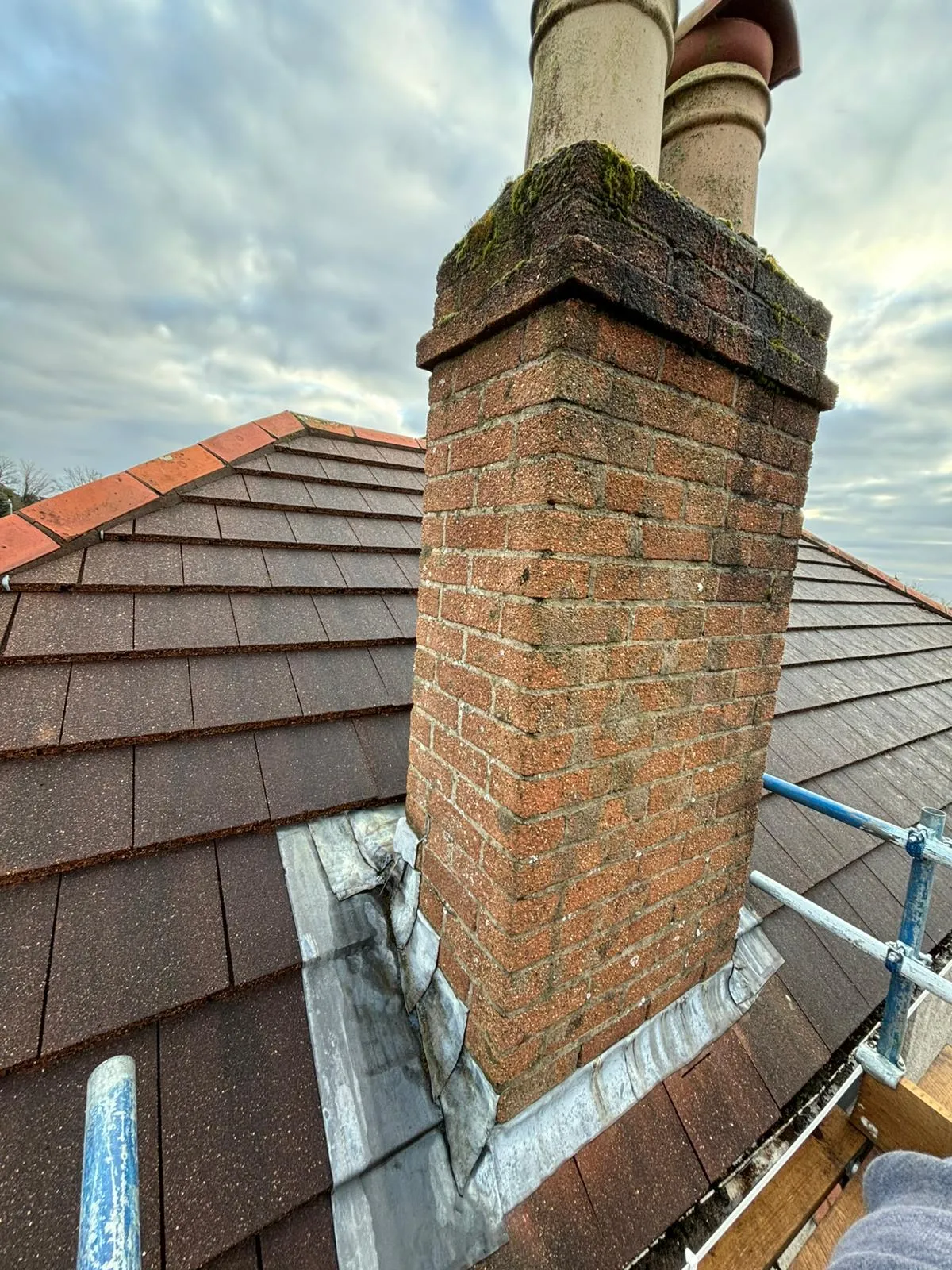 A brick chimney on a tiled roof with lead flashing and scaffolding for repairs
