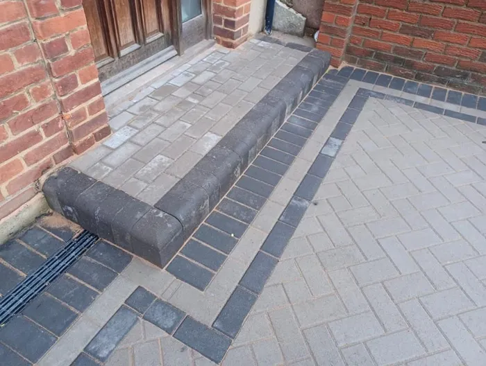 Newly installed light grey block paving with dark grey borders and a step leading to a house entrance, including an integrated drainage channel.