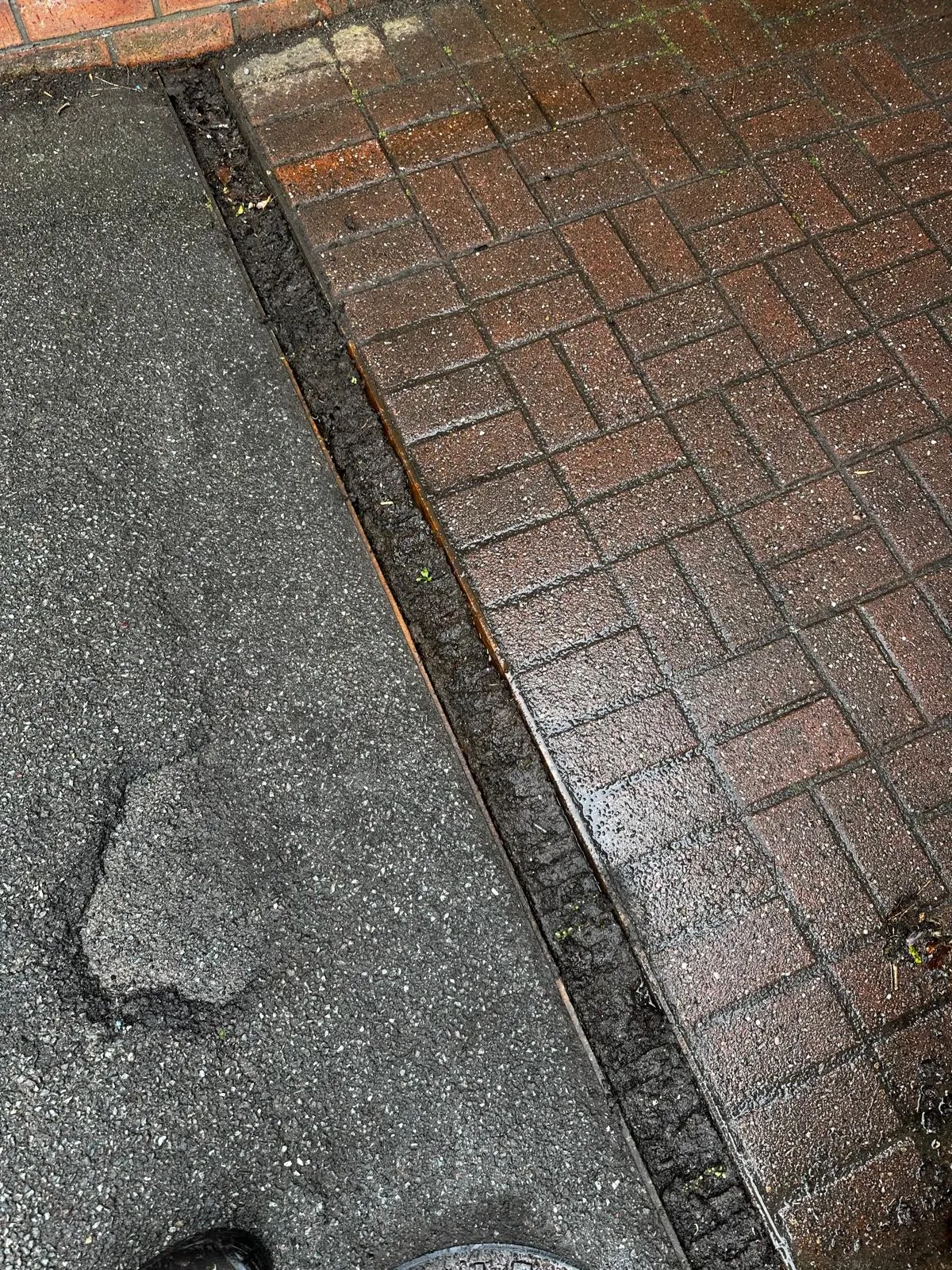 Blocked outdoor drainage gully filled with debris between a tarmac path and brick paving, highlighting the need for professional drain unblocking services in Skipton.