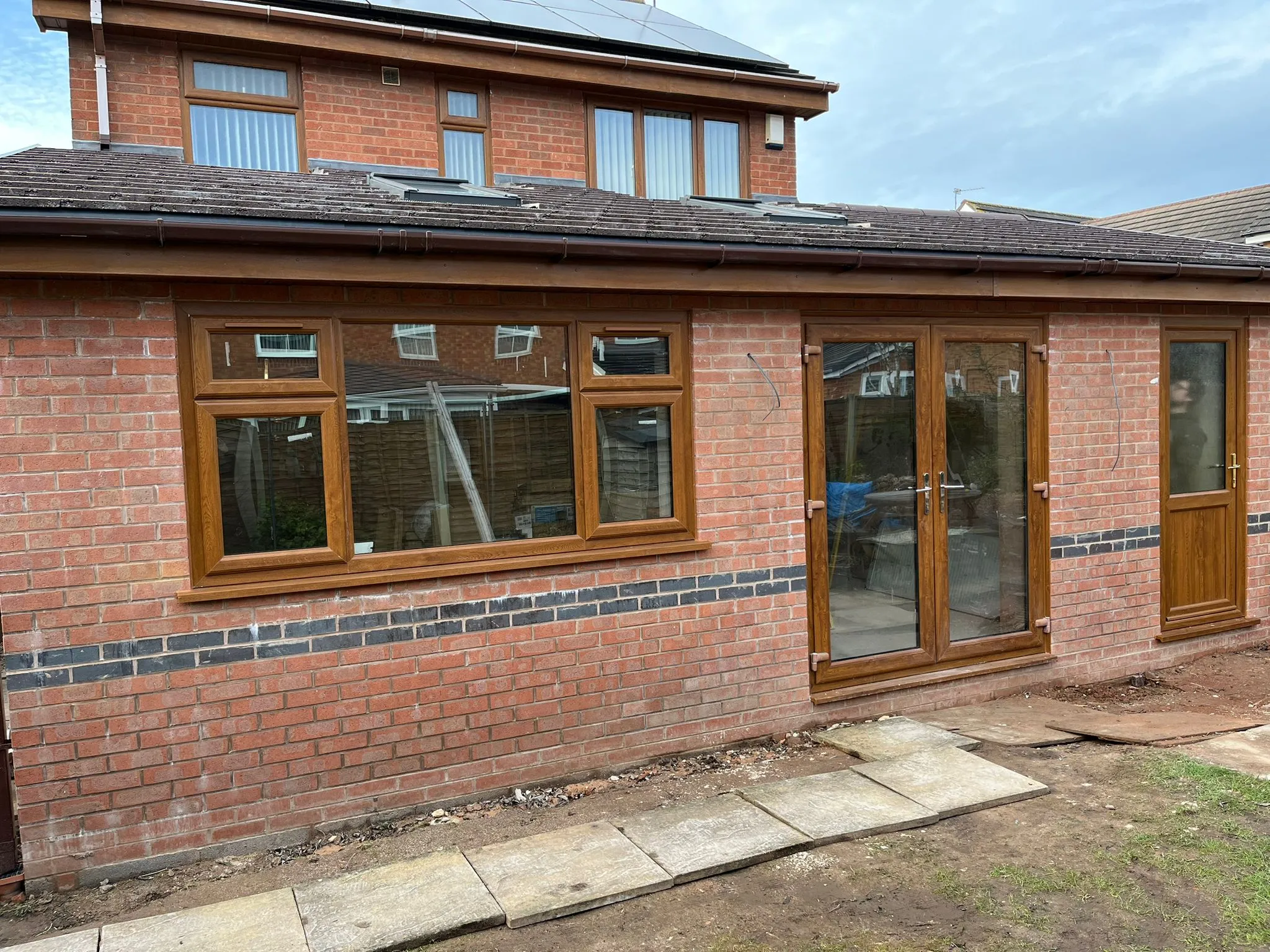 A newly built brick home extension with brown window and door frames, seen from a garden under development.