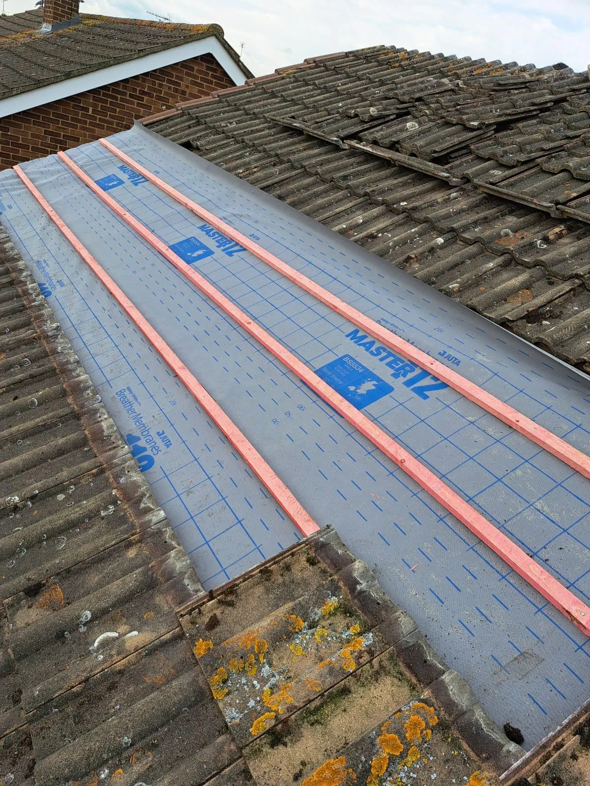 A close-up view of a roof in progress, with half covered in old, moss-covered tiles and the other half with new grey breathable roofing membrane (Juta Master 12) and red wooden battens laid out.