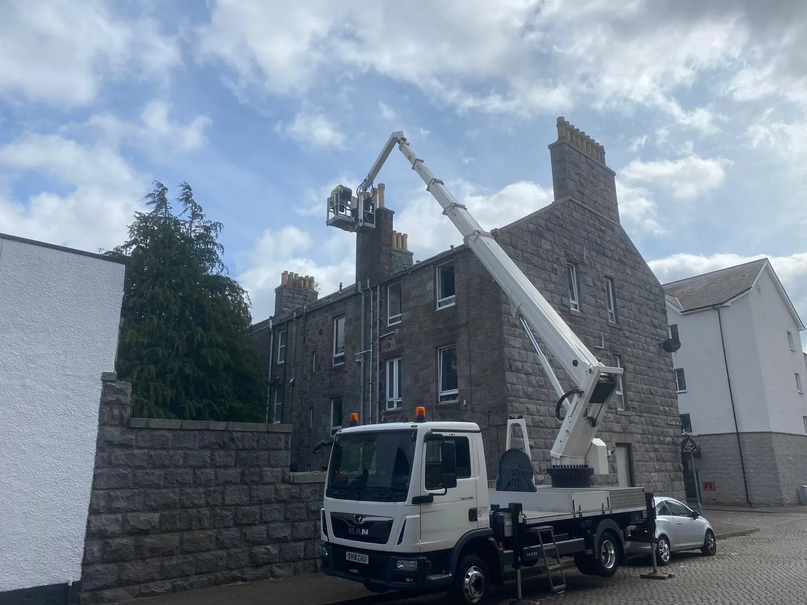 A cherry picker vehicle with a worker in the elevated basket performing chimney repair or inspection on a traditional stone building.