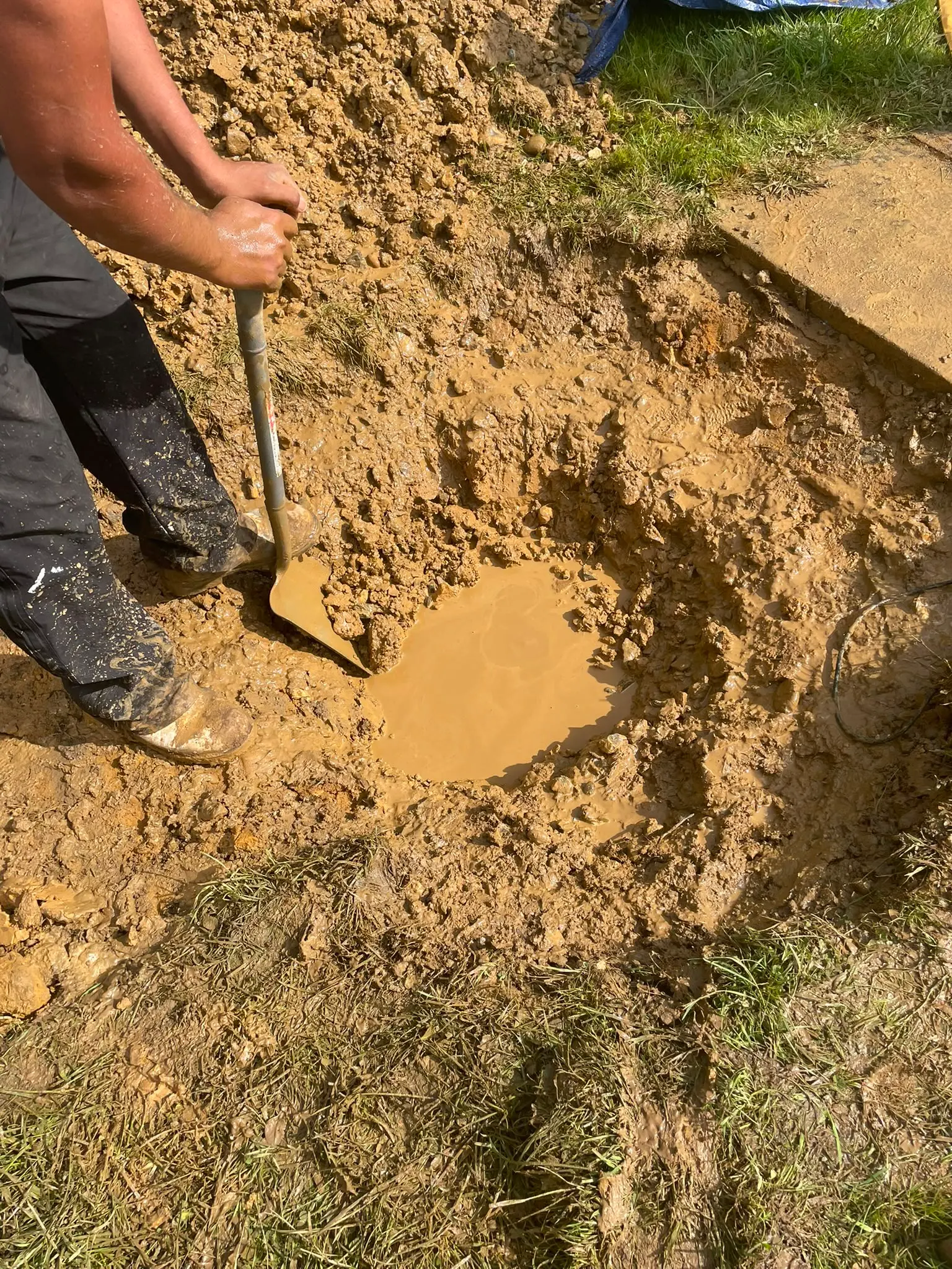 A worker digging a muddy hole in the ground with a shovel for drainage repairs