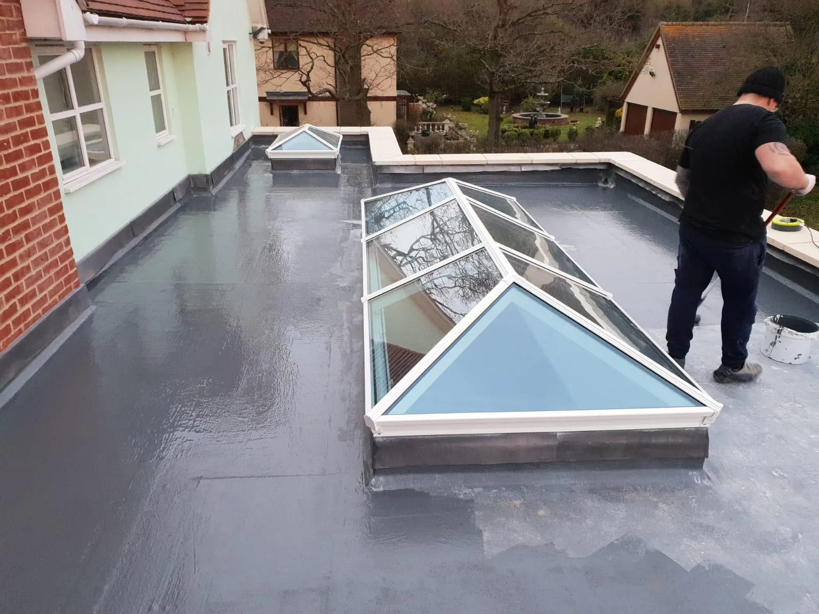 A flat roof installation showing a grey waterproof membrane, two large glass roof lanterns, and a builder working with a roller. The service includes flat roofing and skylight installation.