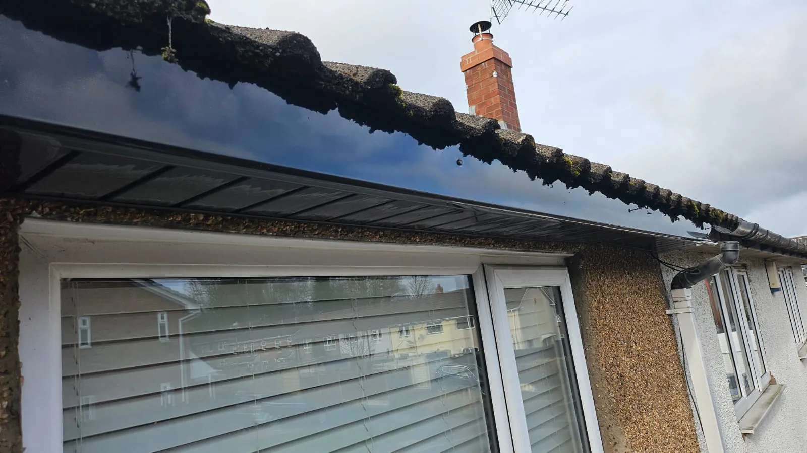 Newly installed black uPVC fascias and soffits beneath a tiled roof with moss, alongside a white window and downpipe.