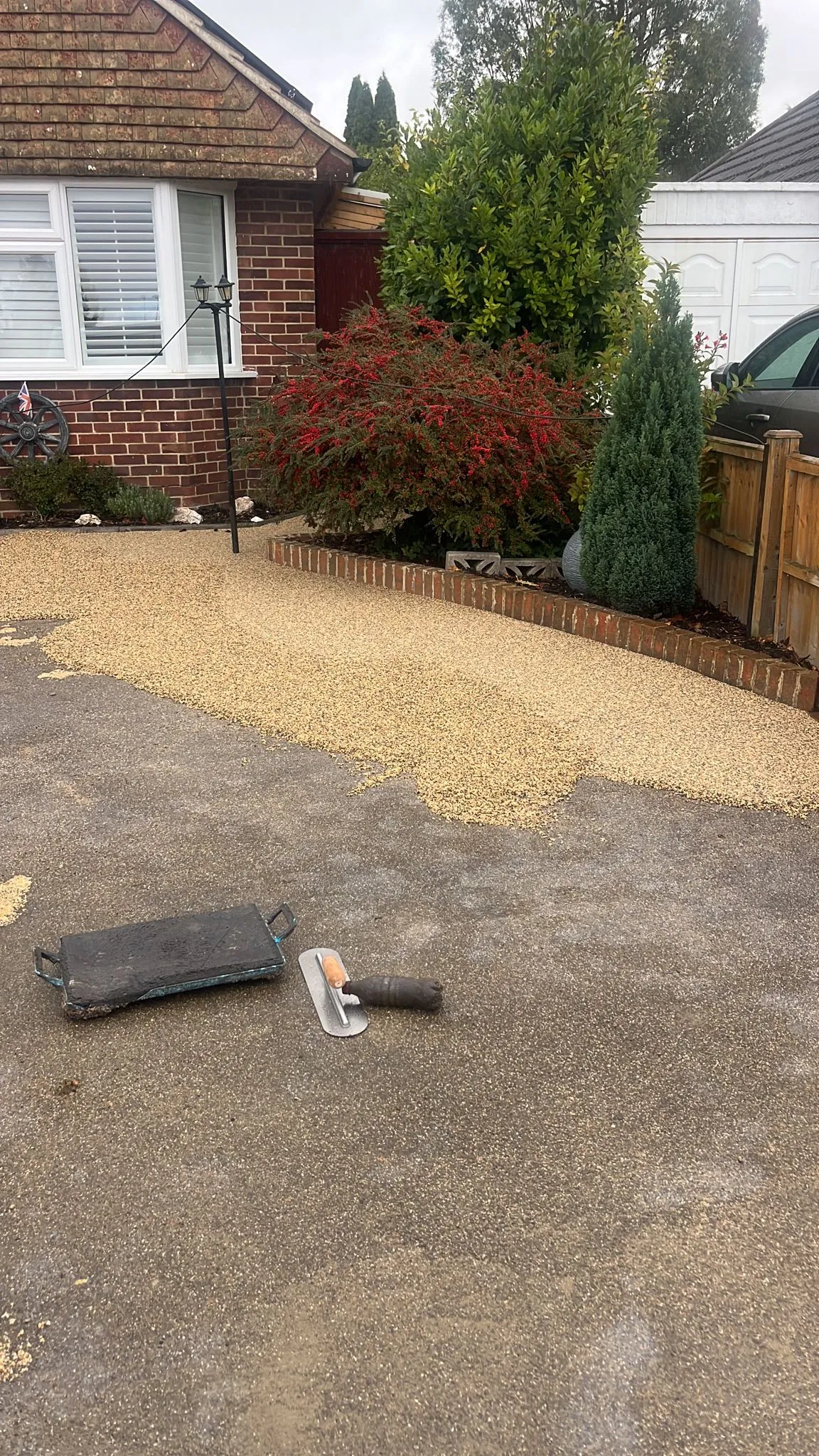 Construction scene of a resin-bound driveway installation, showing a fresh layer of light-coloured aggregate being smoothed over an old tarmac driveway in front of a brick house with garden shrubs. A trowel and float lie on the ground.