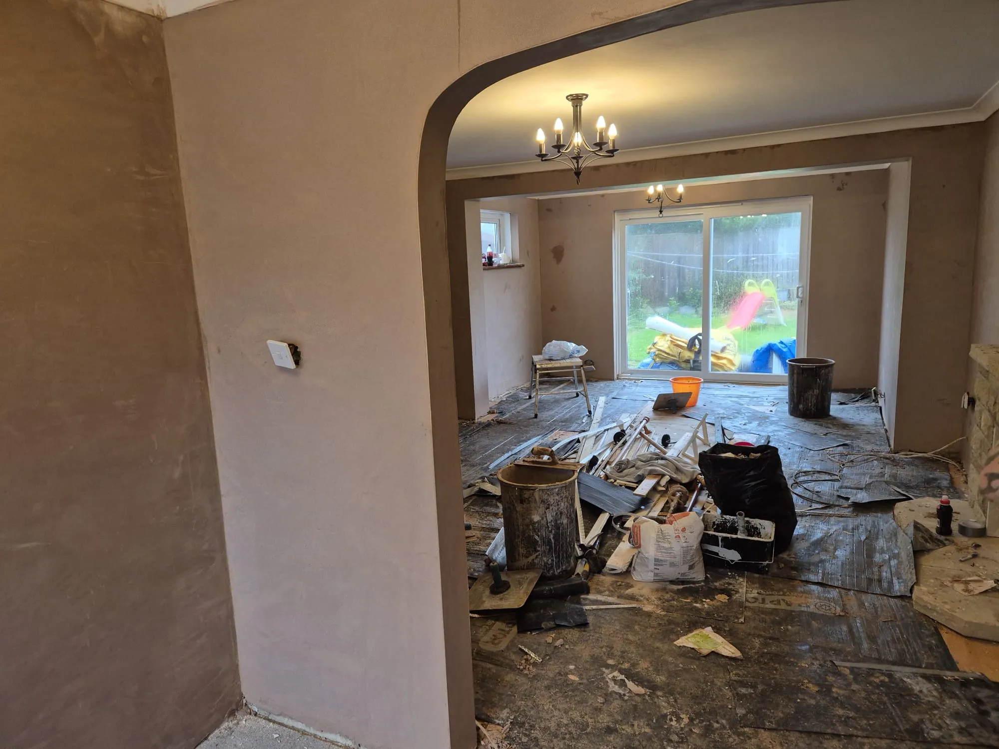 An interior room in mid-refurbishment, showing freshly plastered walls, an archway, and construction materials and debris on the floor, with a view to a garden through sliding doors.