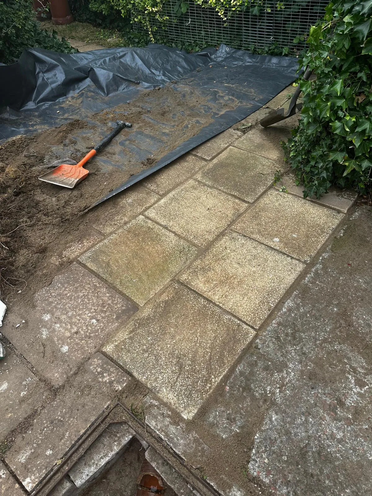 Open drain access point with an exposed terracotta pipe during excavation work in a garden with paving slabs and a shovel.
