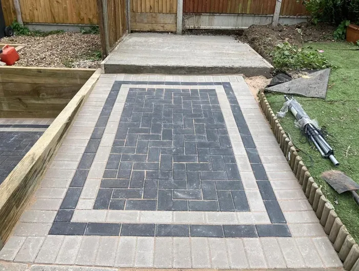 Newly laid block paving path in a garden, with dark grey and light grey blocks, a wooden raised bed, and artificial grass.