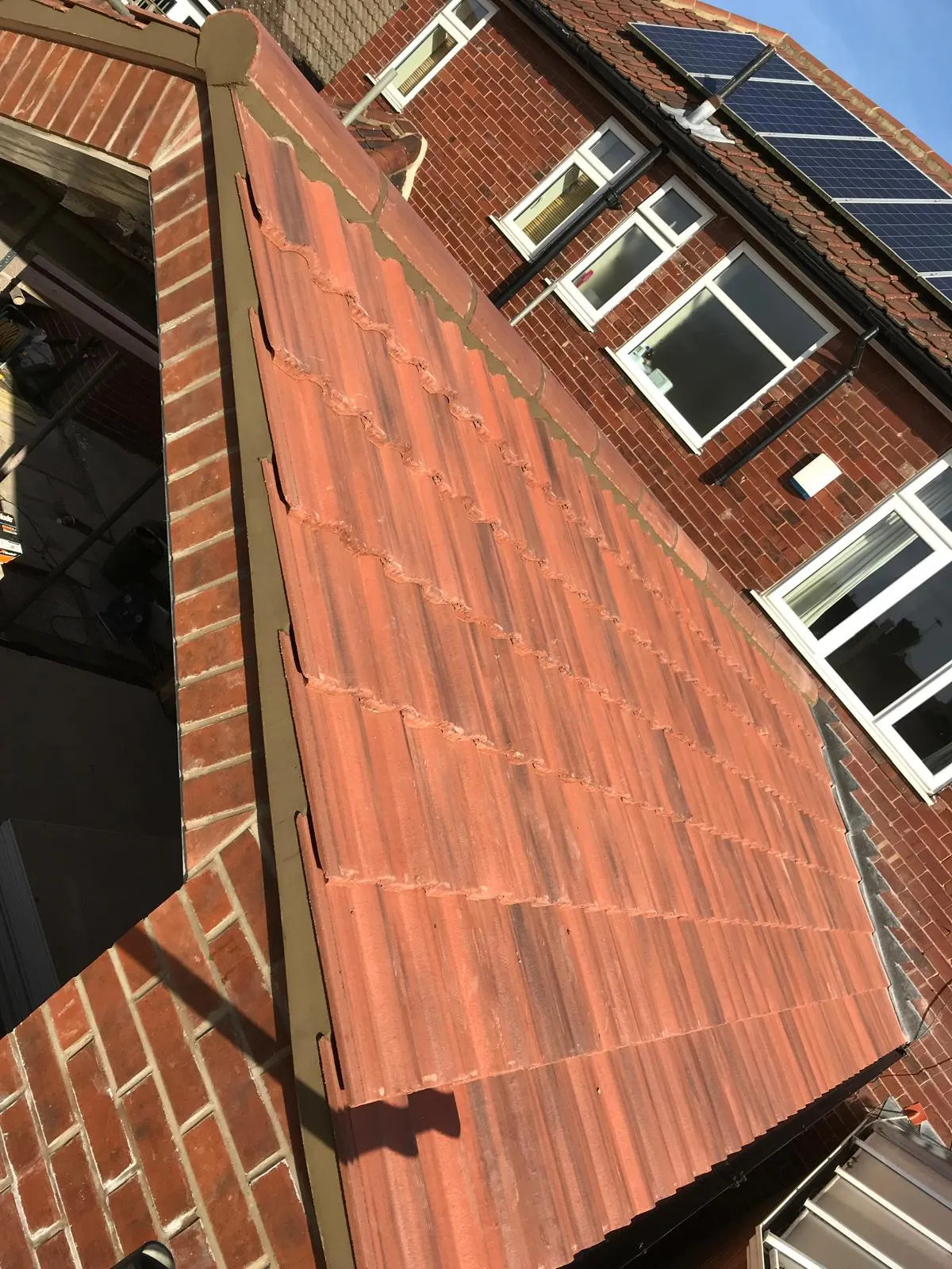 Close-up view of a residential pitched roof with reddish-brown tiles, showing new mortar work on the edges and solar panels on an adjacent roof.