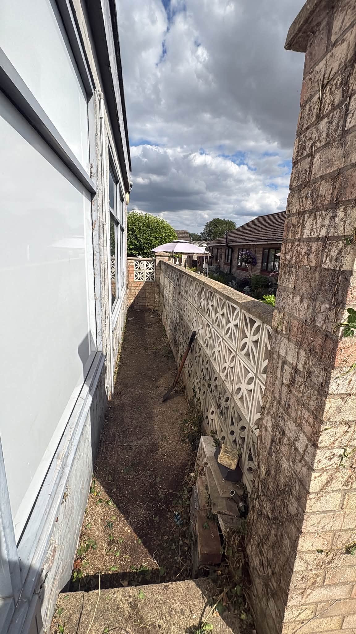 A narrow side path between a house and a decorative breeze block wall, cleared of vegetation down to bare soil, with a shovel leaning against the wall.