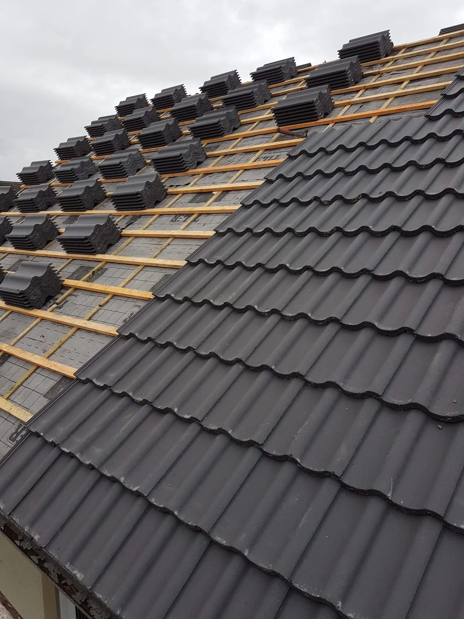 New tiled roof installation in progress showing dark grey concrete tiles being laid over wooden battens and roofing underlay.