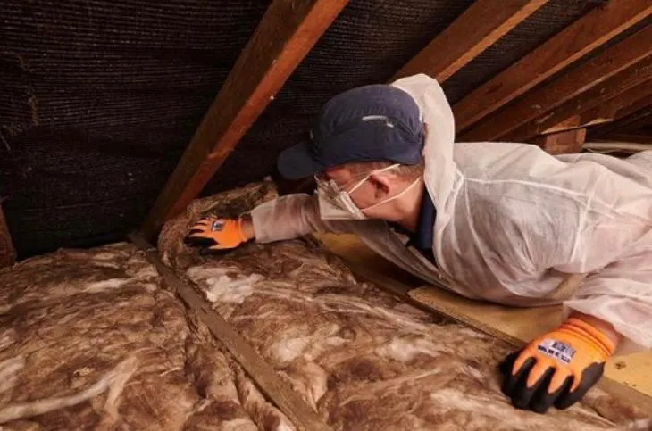 A building contractor in protective suit and mask inspecting loft insulation in an attic.