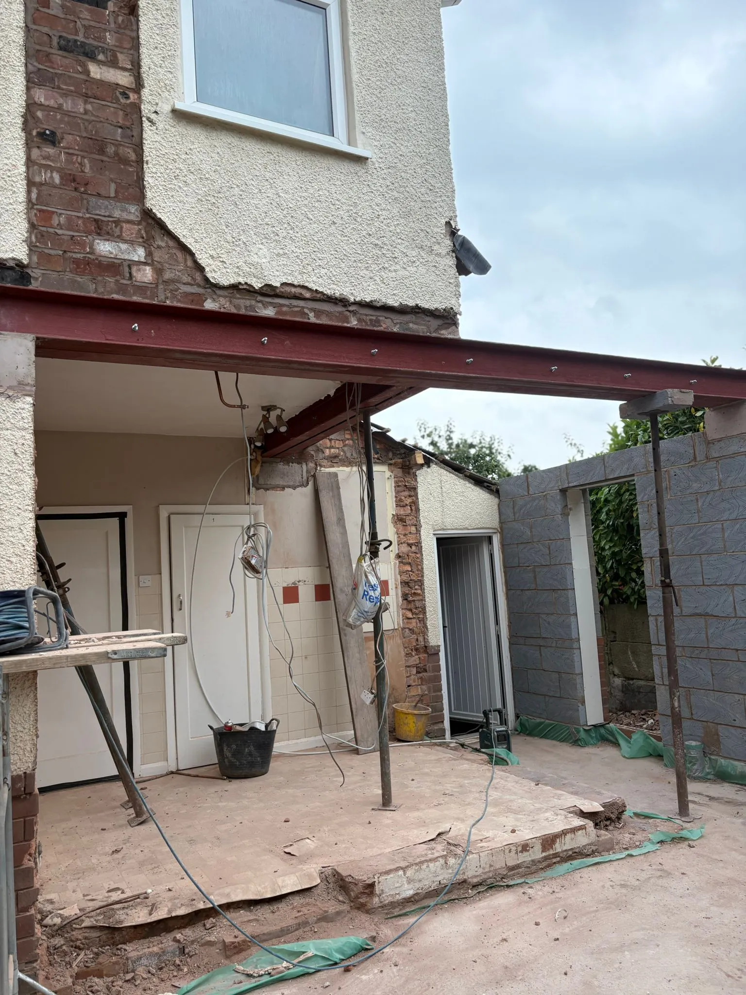 Construction photo of a house extension featuring an exposed brick wall, a newly installed red steel beam for structural support, and the initial stages of new blockwork walls under a cloudy sky.