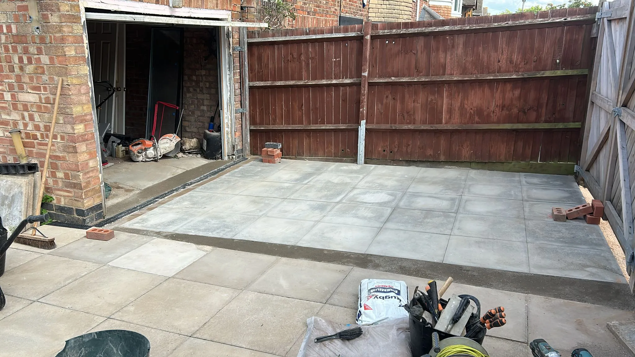 Newly laid grey patio slabs in a garden area, with a timber fence and a drainage channel, during construction.