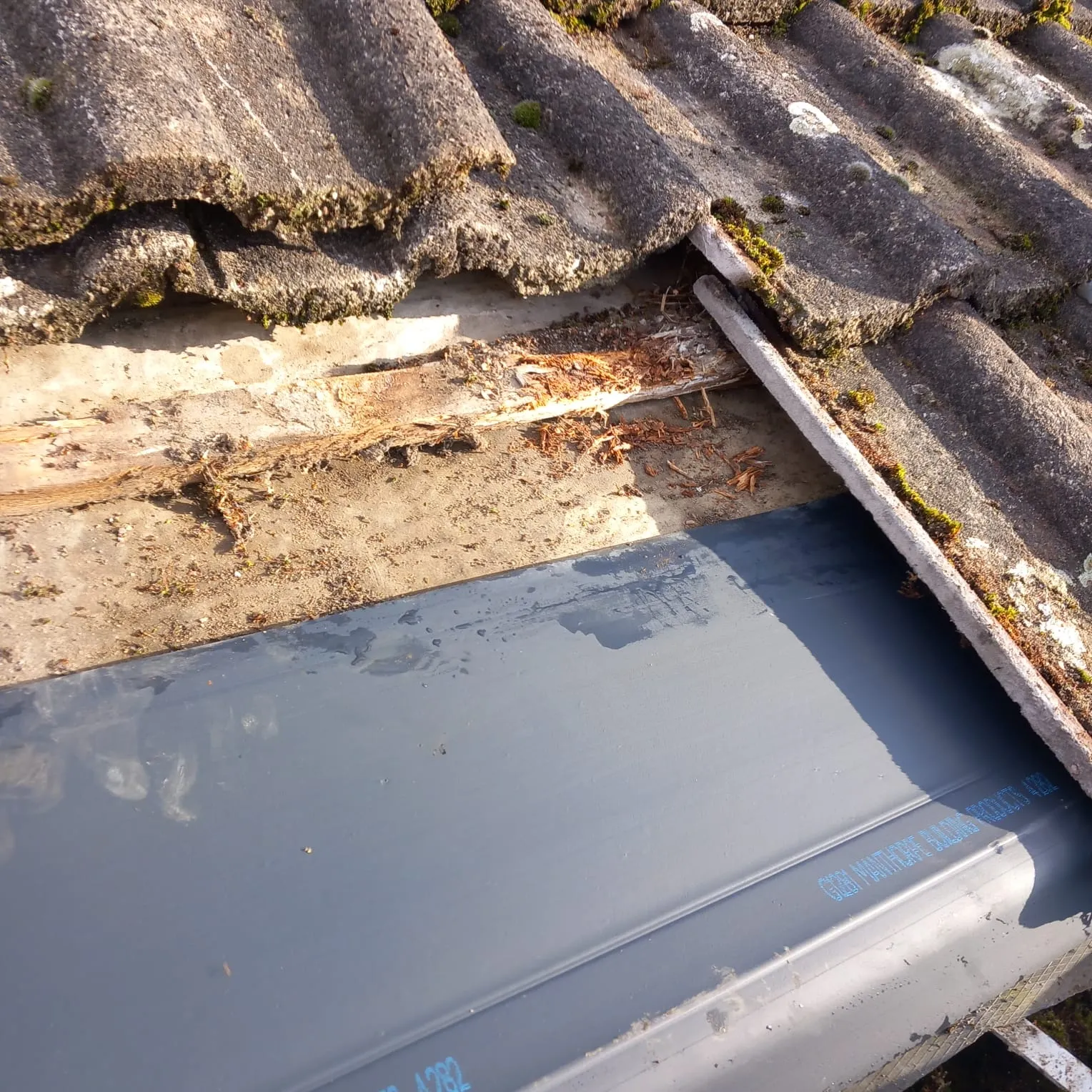 Close-up of a damaged tiled roof with significant moss, exposed wooden battens, and a new grey gutter lining being fitted or repaired.