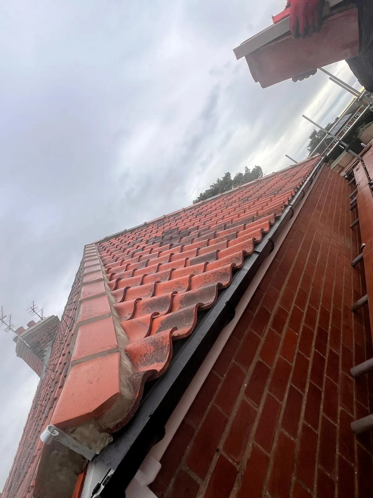 A close-up view of a roofer's gloved hands holding a roof tile, positioned above a pitched roof with red/orange concrete or clay tiles, a brick wall, and scaffolding visible on the right, against an overcast sky.