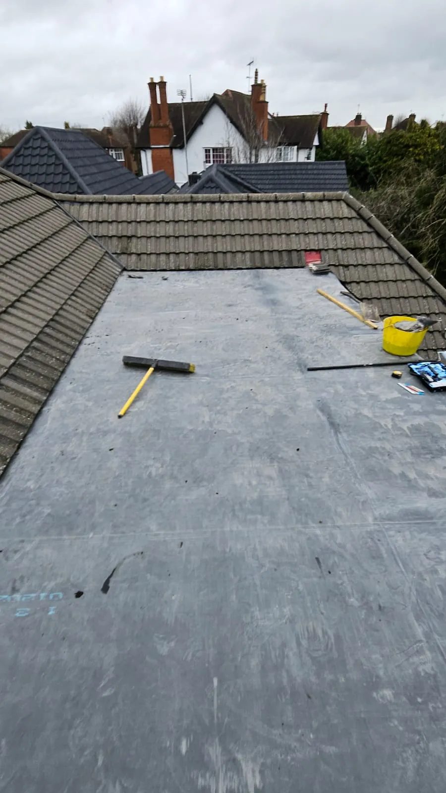 View of a flat roof undergoing installation or repair with a broom and tools, bordered by tiled roofs and houses.