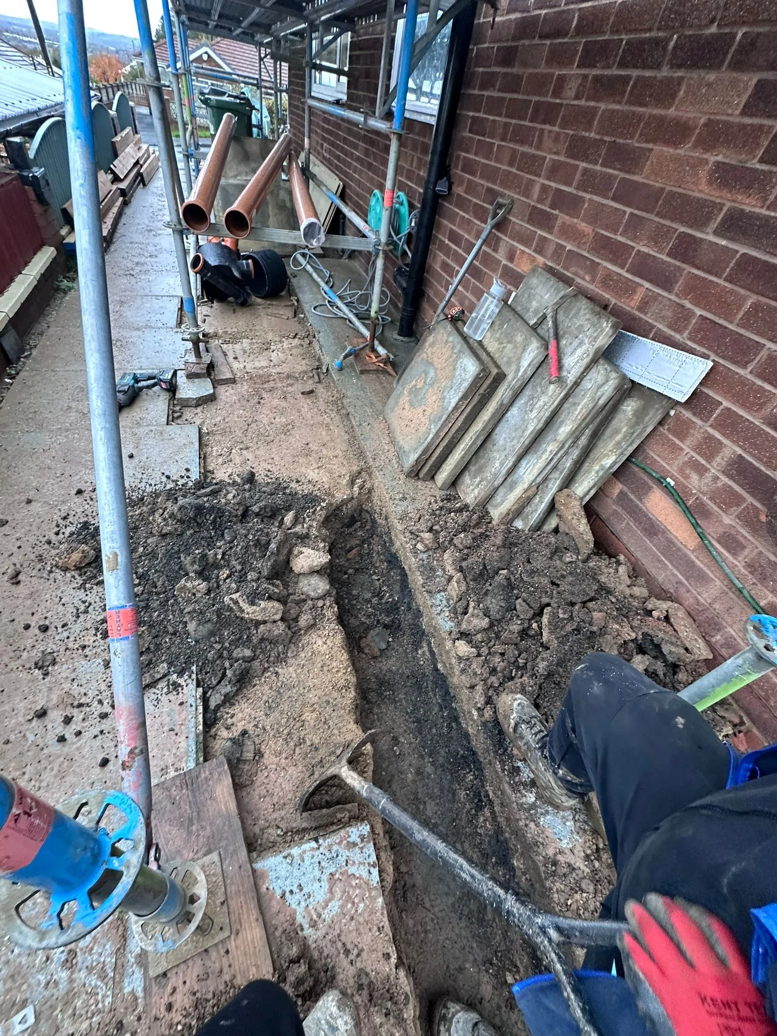 Professional excavation for drain repairs showing a trench, stacked paving slabs, and new drainage pipes at a residential property.