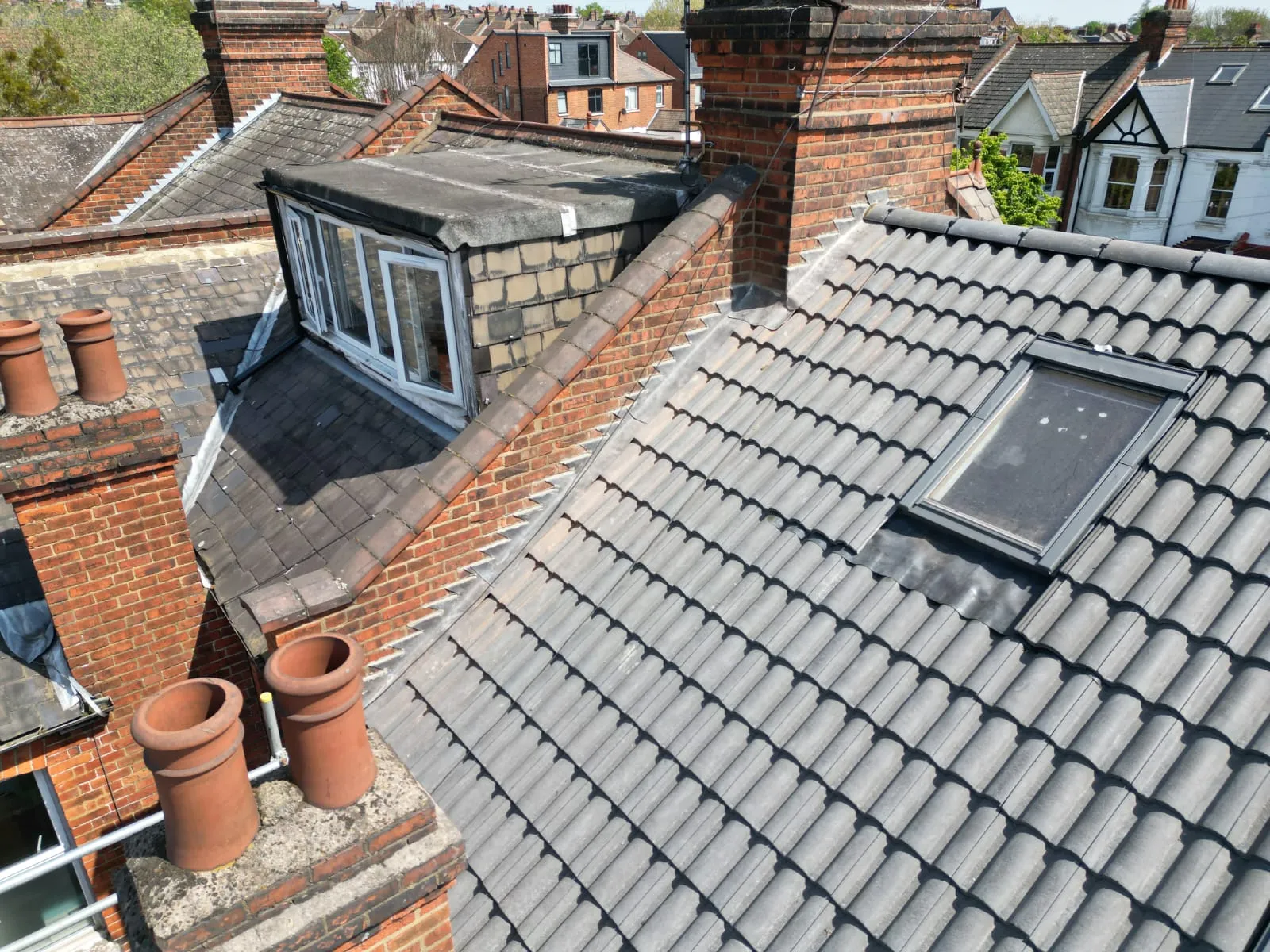 A high-angle view of multiple residential roofs with grey tiles on pitched sections, a flat roof dormer window, and brick chimneys, showcasing different roofing elements.