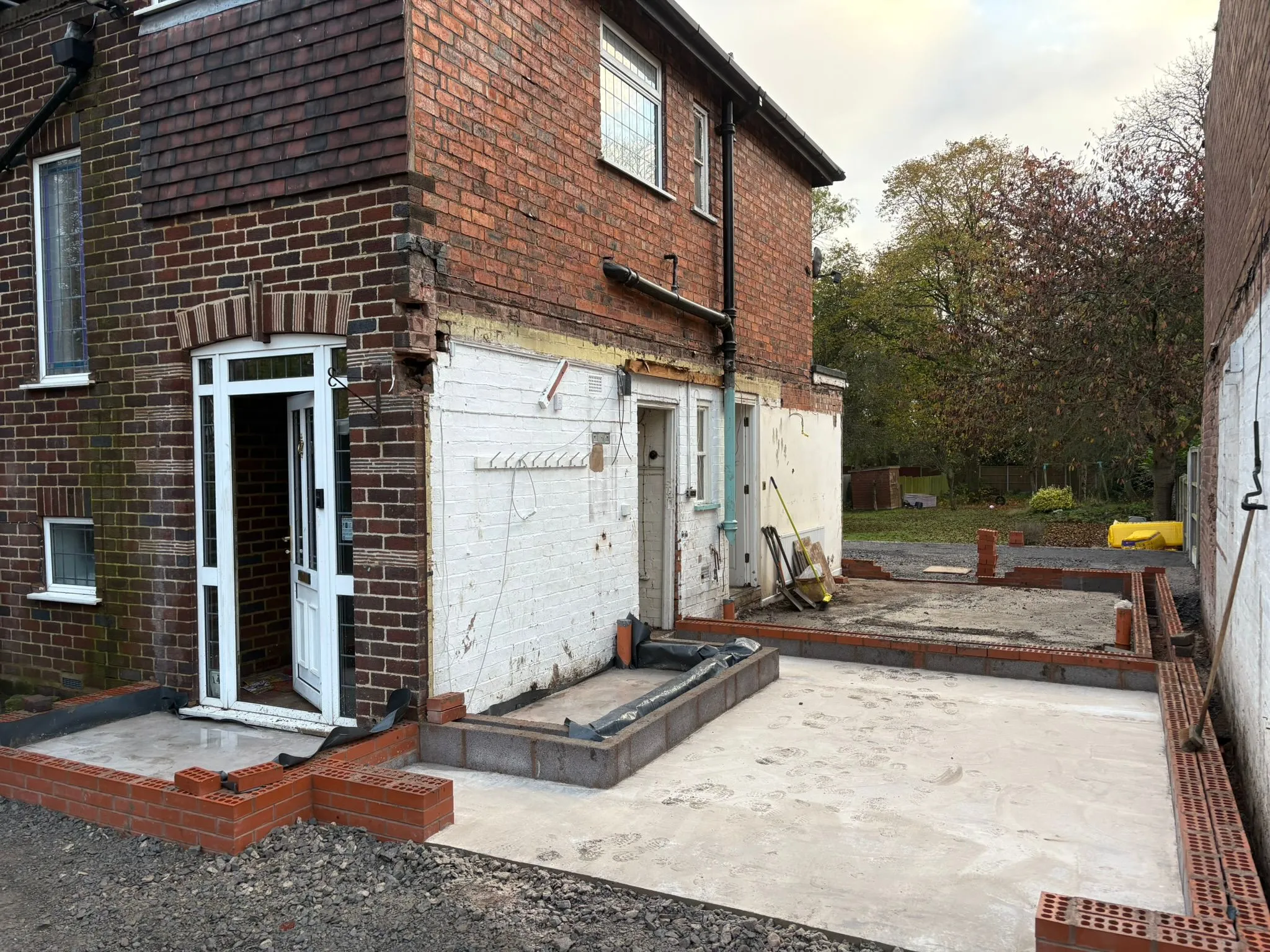 A house undergoing an extension build, with concrete foundations and the first courses of brickwork for the new structure visible.