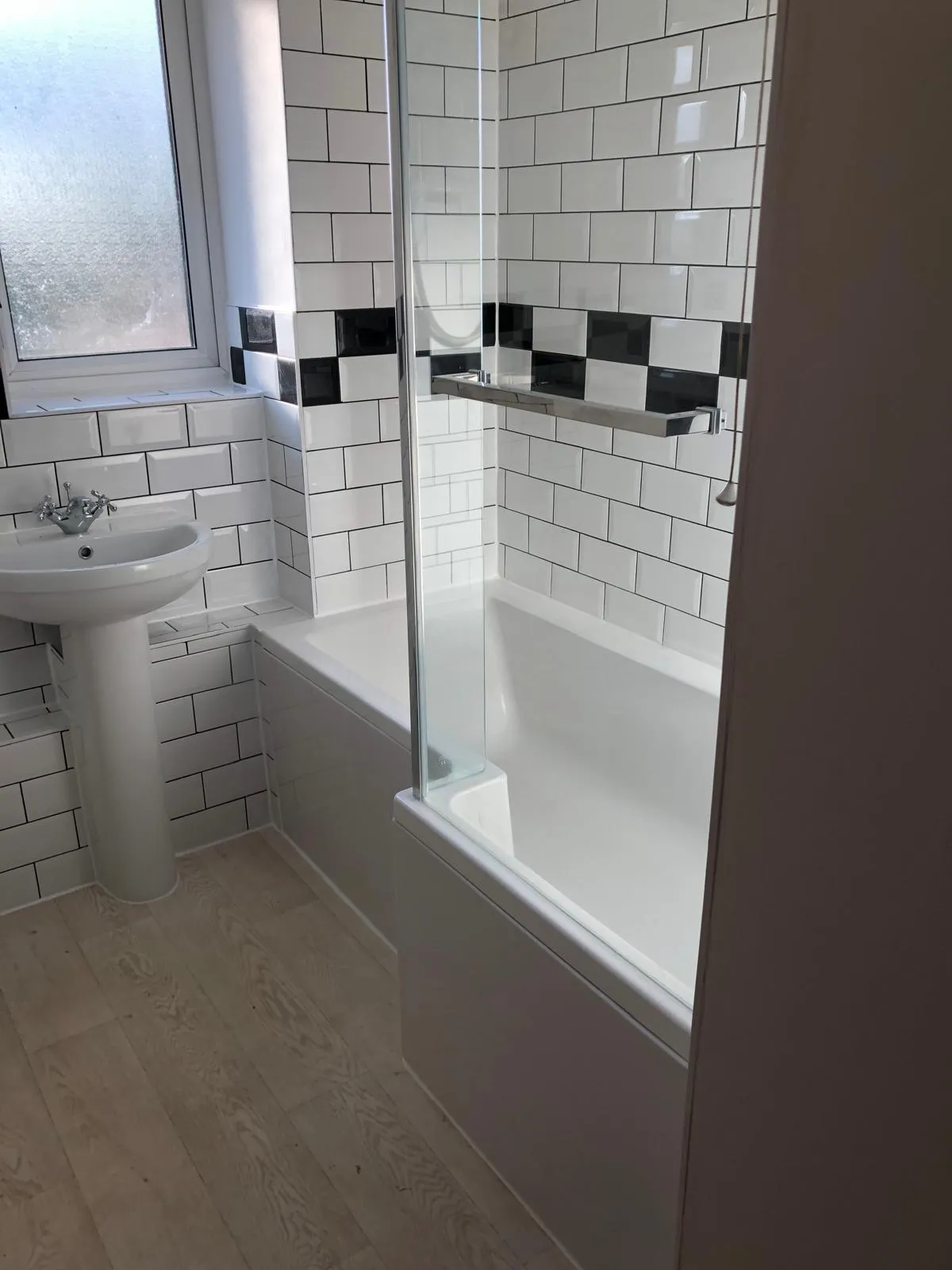 A freshly renovated bathroom with white subway tiles, a black patterned border, a white pedestal sink, and a modern bathtub with a glass screen.