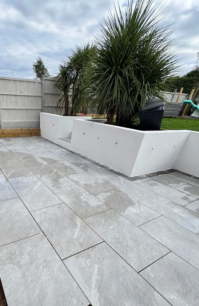 Modern garden with newly laid grey patio tiles, a white rendered raised planter with Cordyline Australis palm trees, and a grey concrete fence.