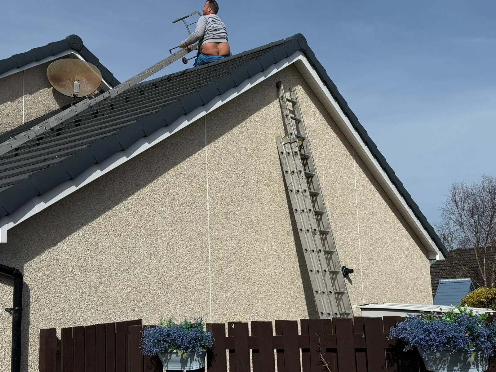 An experienced roofer on a modern tiled pitched roof, using a smaller access ladder to move across the roof, with a satellite dish and main access ladder visible on the side of the house under a clear sky, ready for roofing work or repairs.
