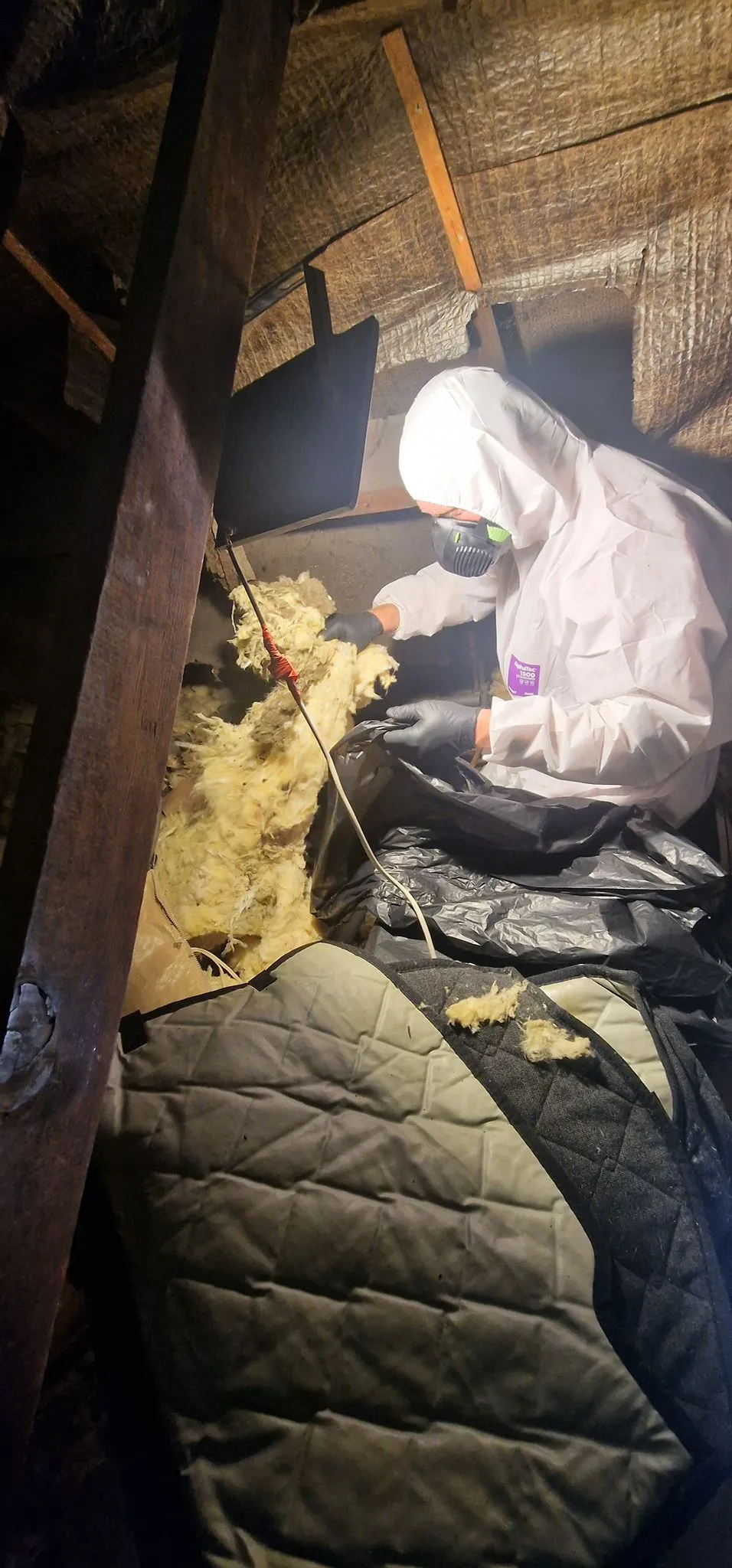 A pest control technician in a white hazmat suit and respirator mask removing old insulation from a dark loft space during a sanitisation service.