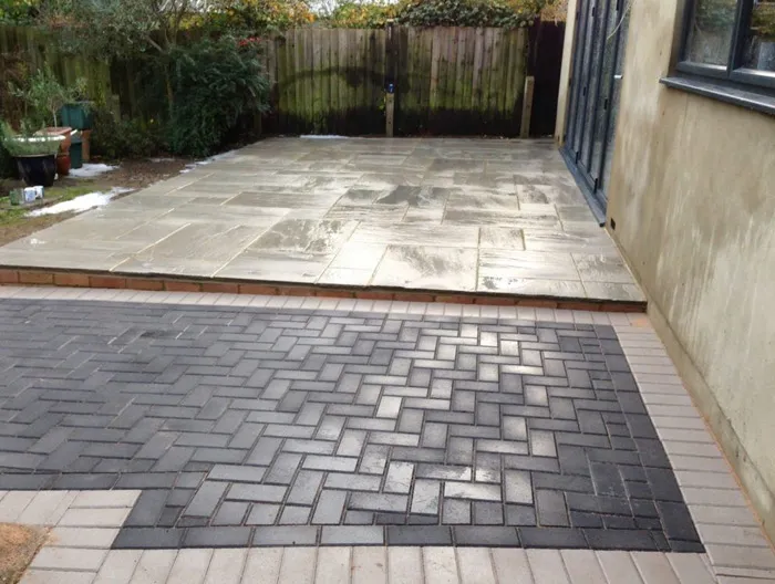 Newly installed dark and light grey block paved driveway with a herringbone pattern, leading to a raised light-coloured natural stone slab patio with a brick step, next to a house with bi-fold doors.