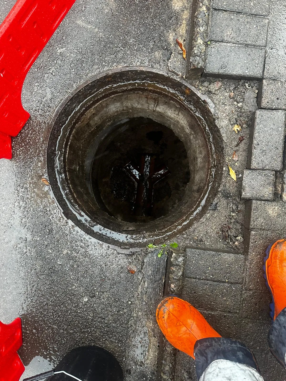 Top-down view of an open manhole on a wet, paved area, with a worker's orange boots and red safety barrier partially visible.